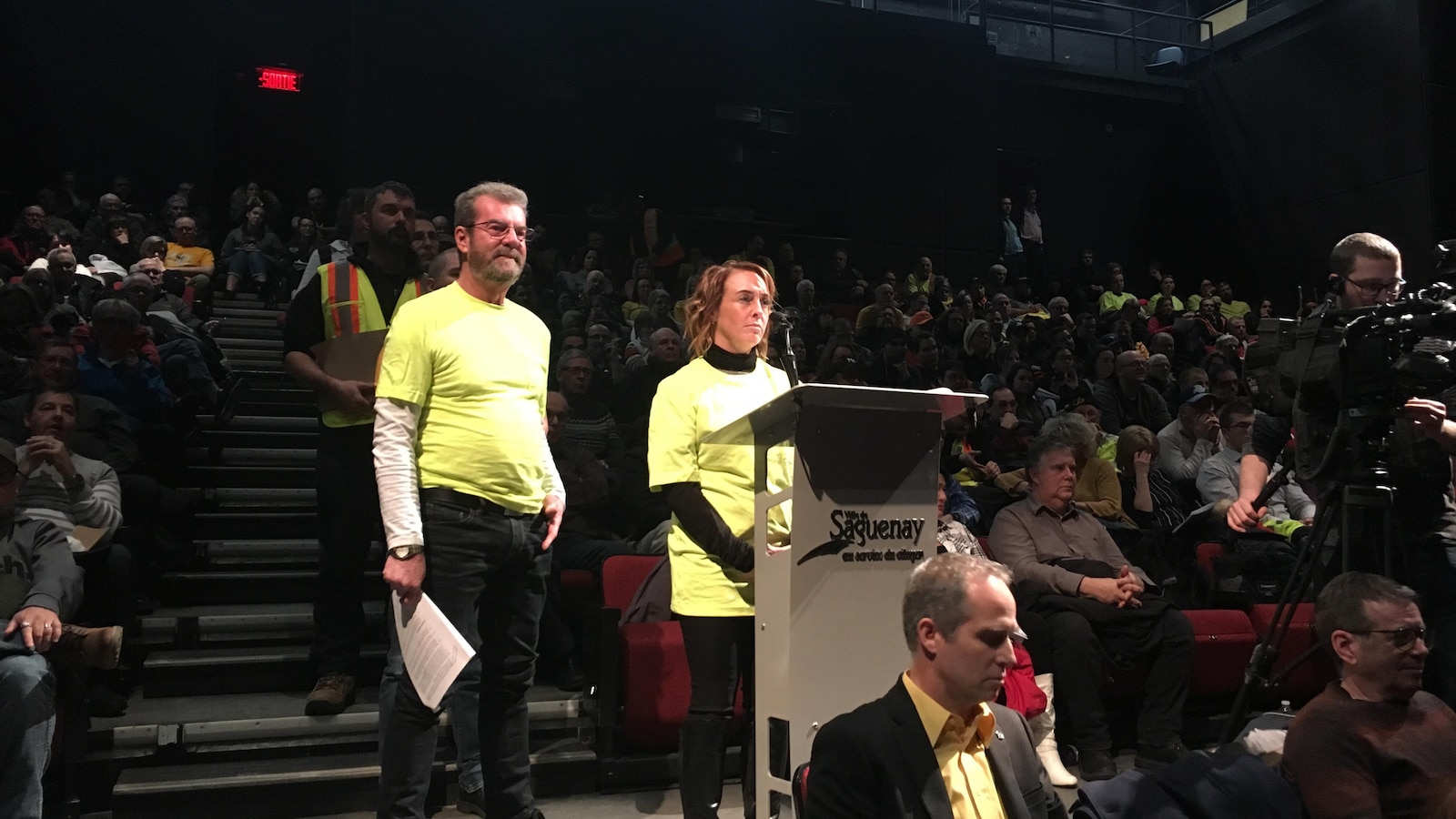 Un homme et une femme sont debout devant un lutrin. Ils attendent de poser une question à la mairesse.