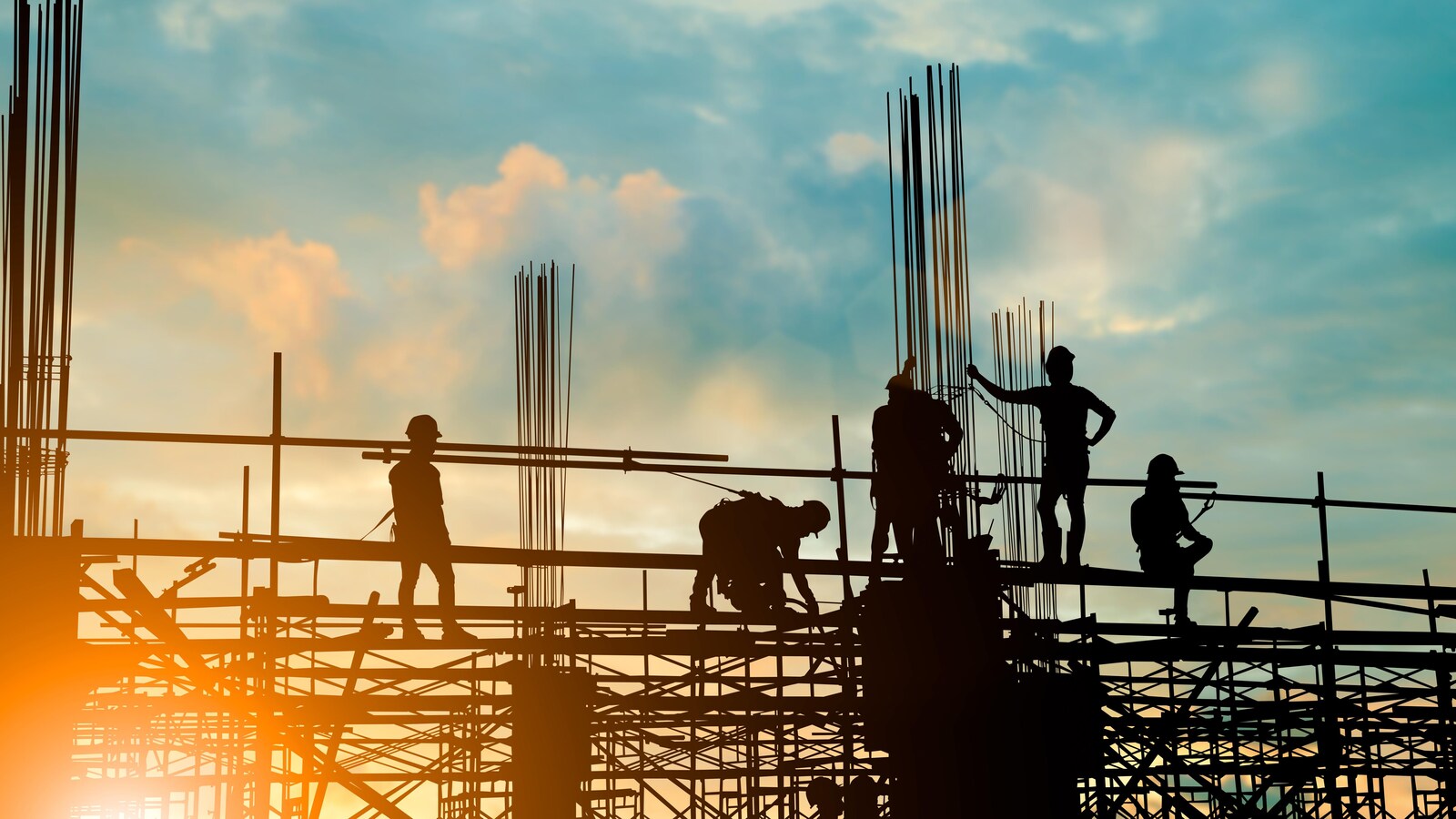 Les silhouettes de personnes sur une partie surélevée d'un chantier de construction sont vues lors d'un coucher ou lever du soleil. Ces personnes portent des casques protecteurs.