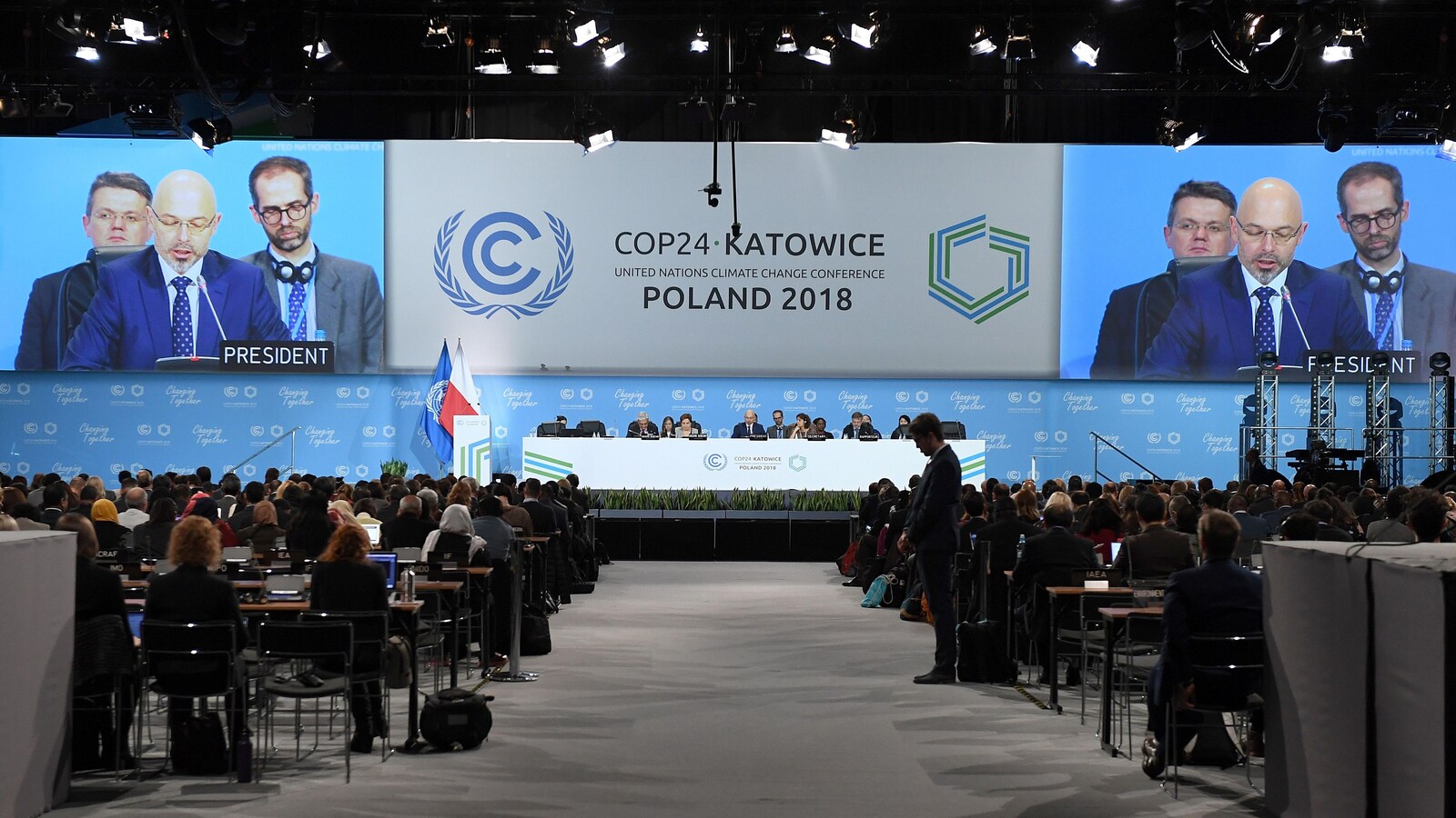 D'énormes écrans diffusent le discours d'un homme assis à une longue table sur une estrade à l'intérieur d'un stade où sont réunis plusieurs diplomates en Pologne.
