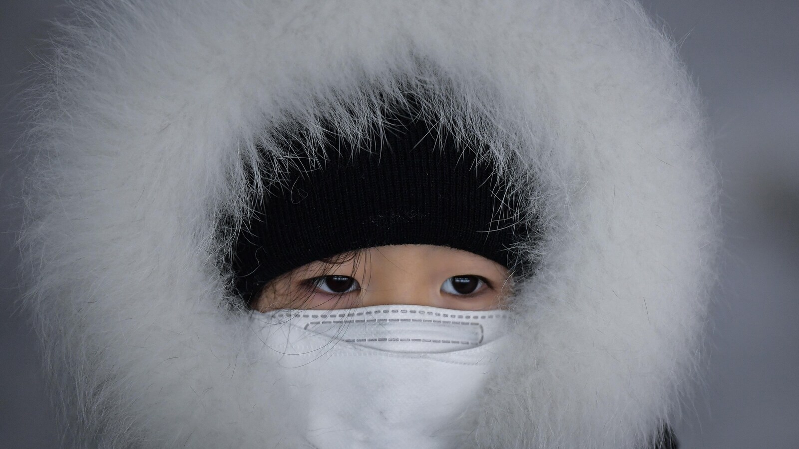 Un enfant la tête couverte d'un capuchon de fourrure et d'une tuque avec la bouche couverte d'un masque blanc assiste à une descente de luge à Pyeongchang, en Corée du Sud.