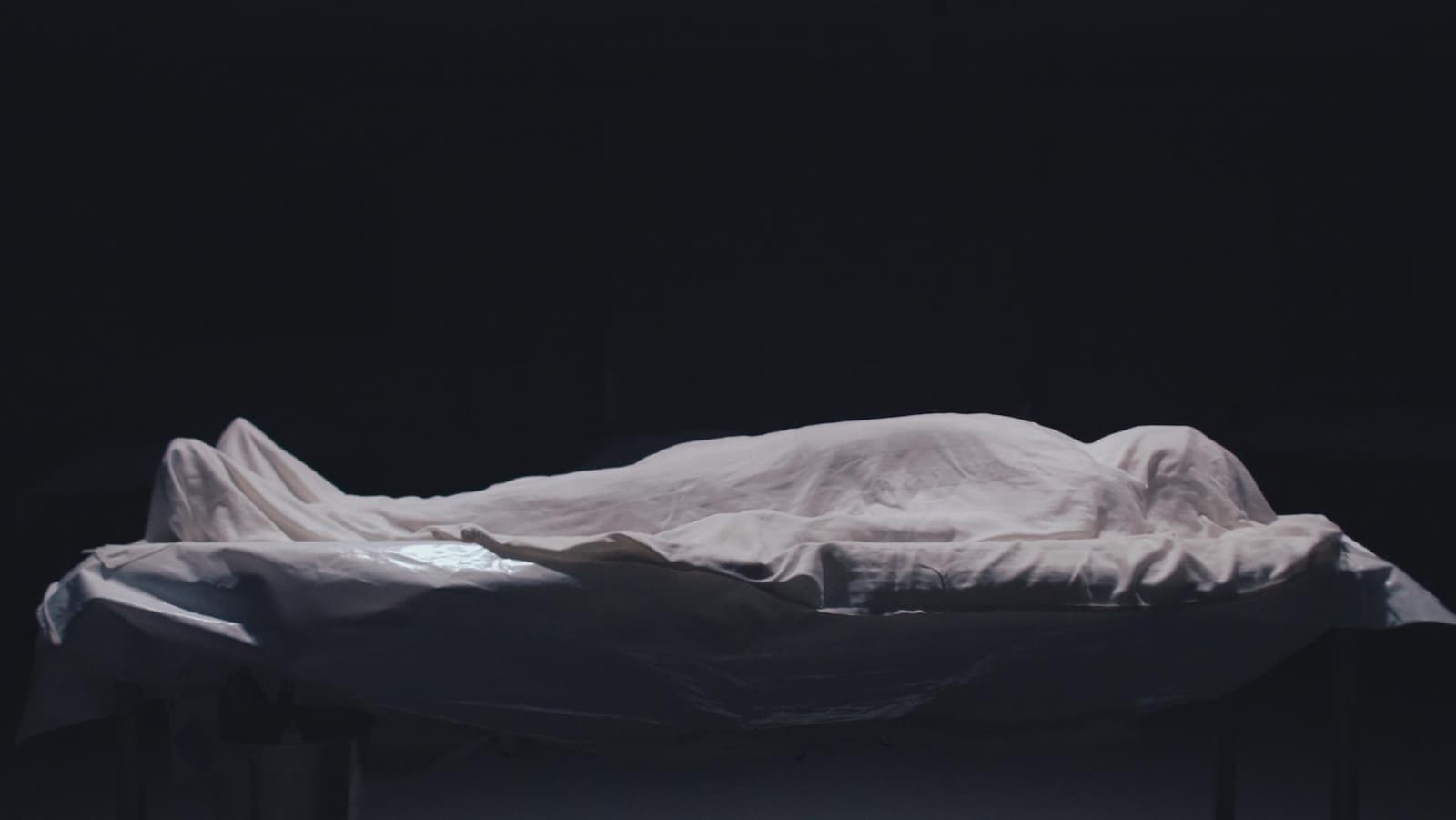 Un drap blanc recouvre un cadavre placé sur une table. Une lumière blanche l'éclaire de haut.
