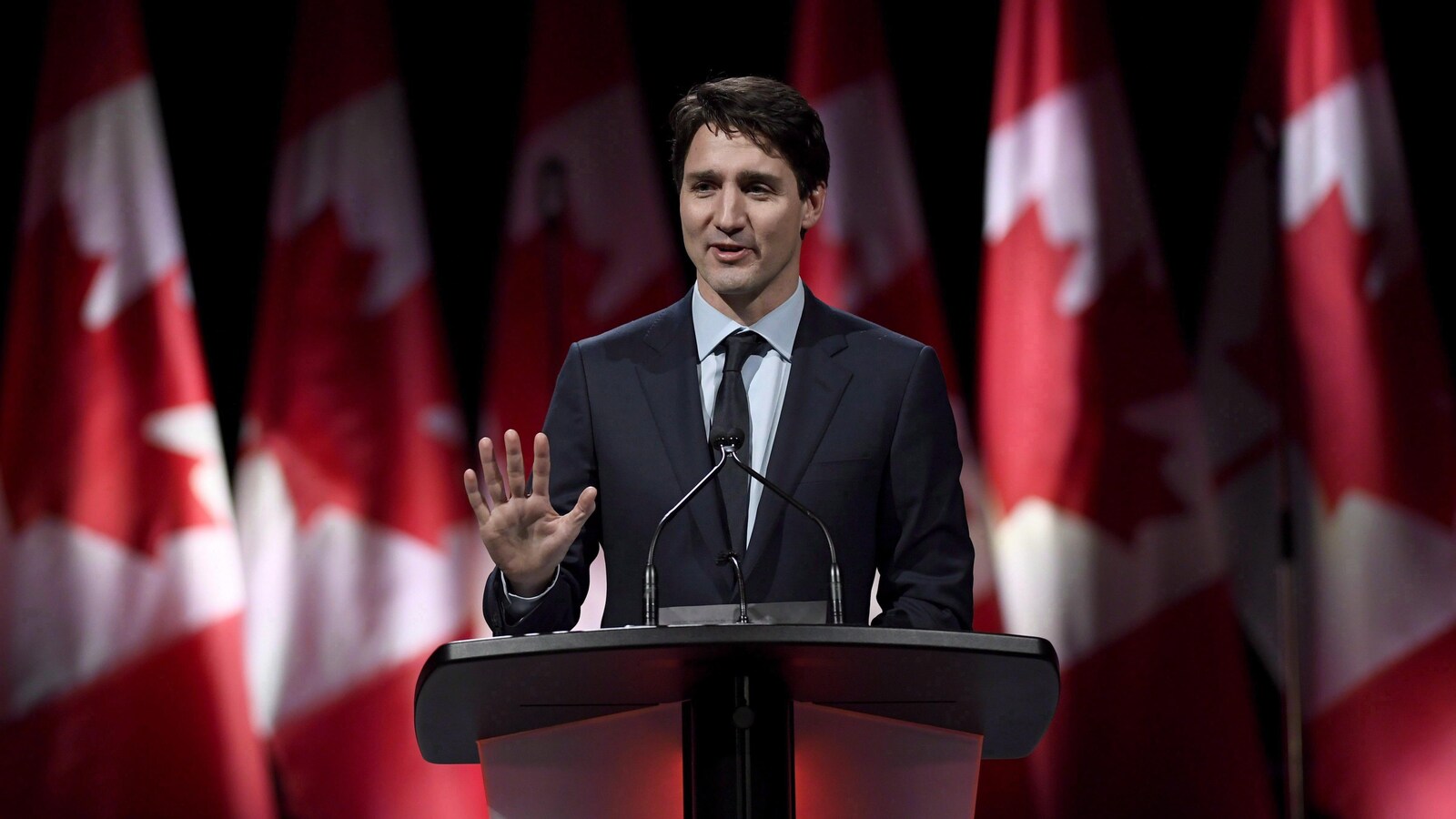 Justin Trudeau sourit, levant la main, devant une série de drapeaux du Canada.