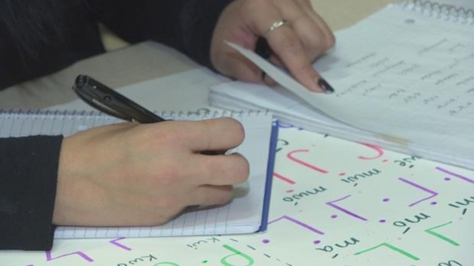 Des mains de femmes écrivent et tournent la page d'un cahier où il y a des lettres cries.