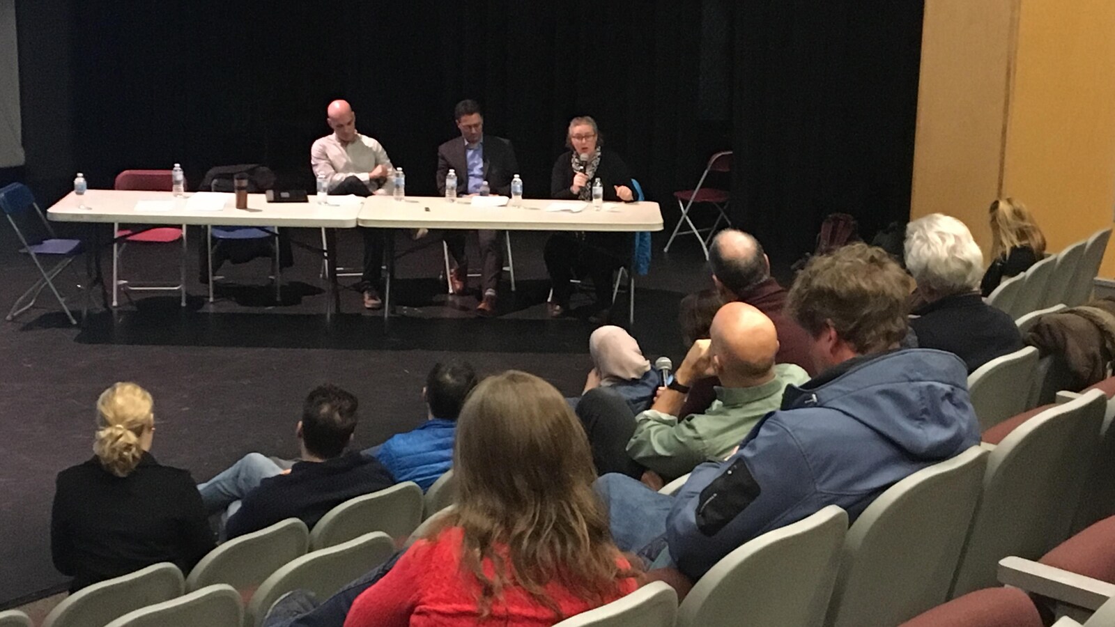 Des parents de dos, dans un auditorium, qui écoutent trois personnes sur scène, assises à une table.