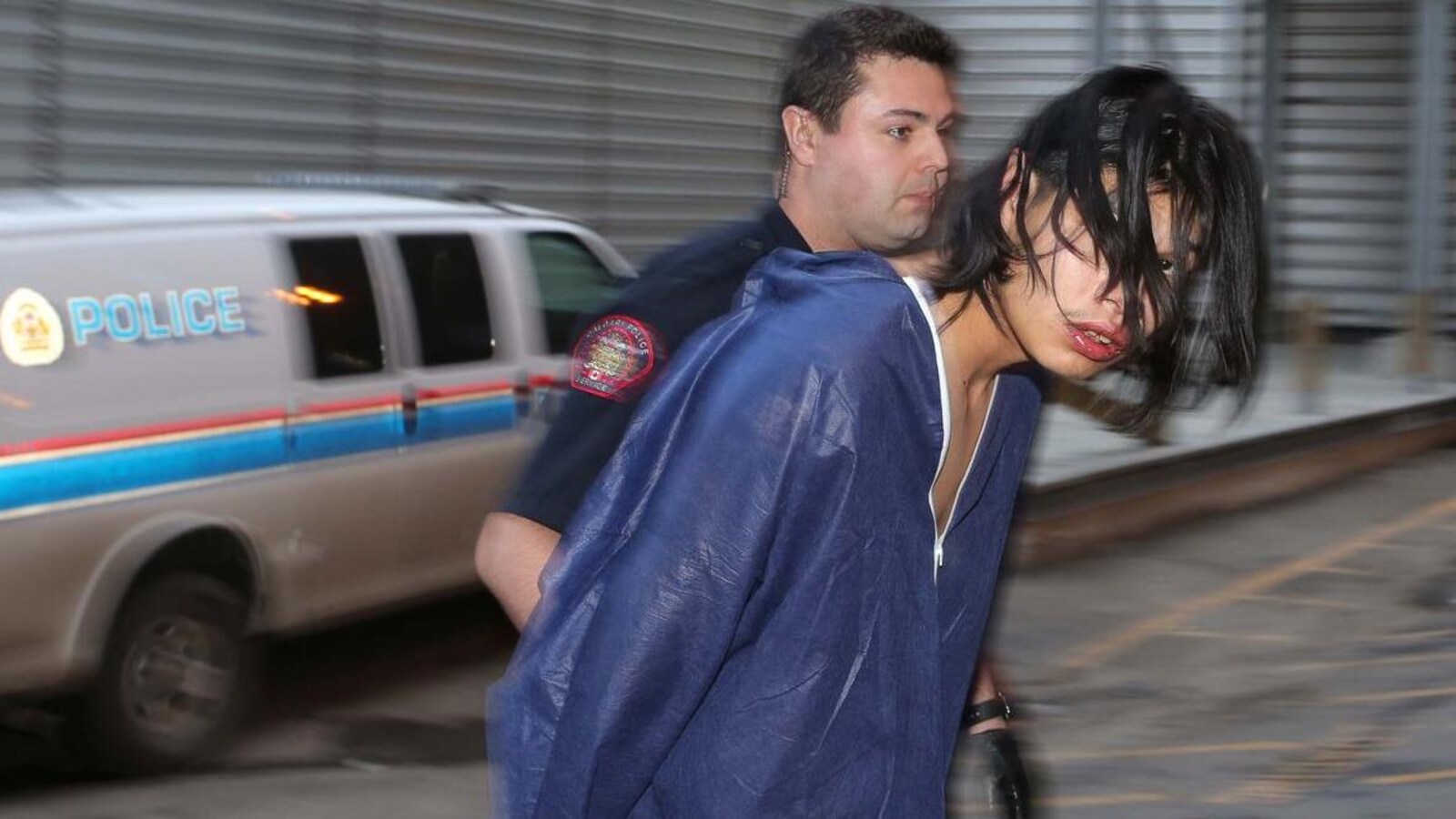 Vue sur un homme avec des cheveux noirs dans le visage et vêtu d'un habit bleu. Il est escorté par un policier.