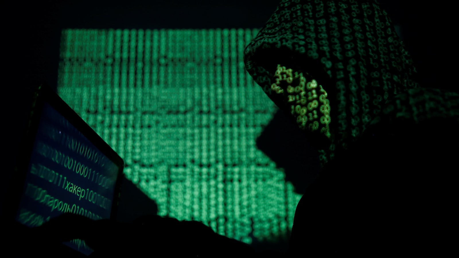 Un homme portant un capuchon, dont le visage est remplacé par du code informatique, est assis devant un écran où l'on voit aussi du code informatique.