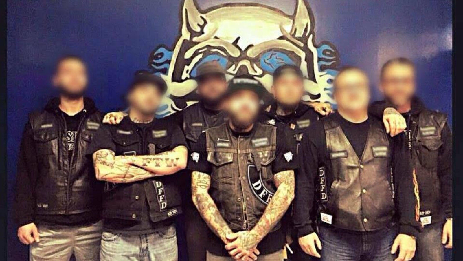 Les membres du groupe de motards Darksiders North Shore prennent la pose devant une affiche du groupe. Leurs visages sont brouillés.