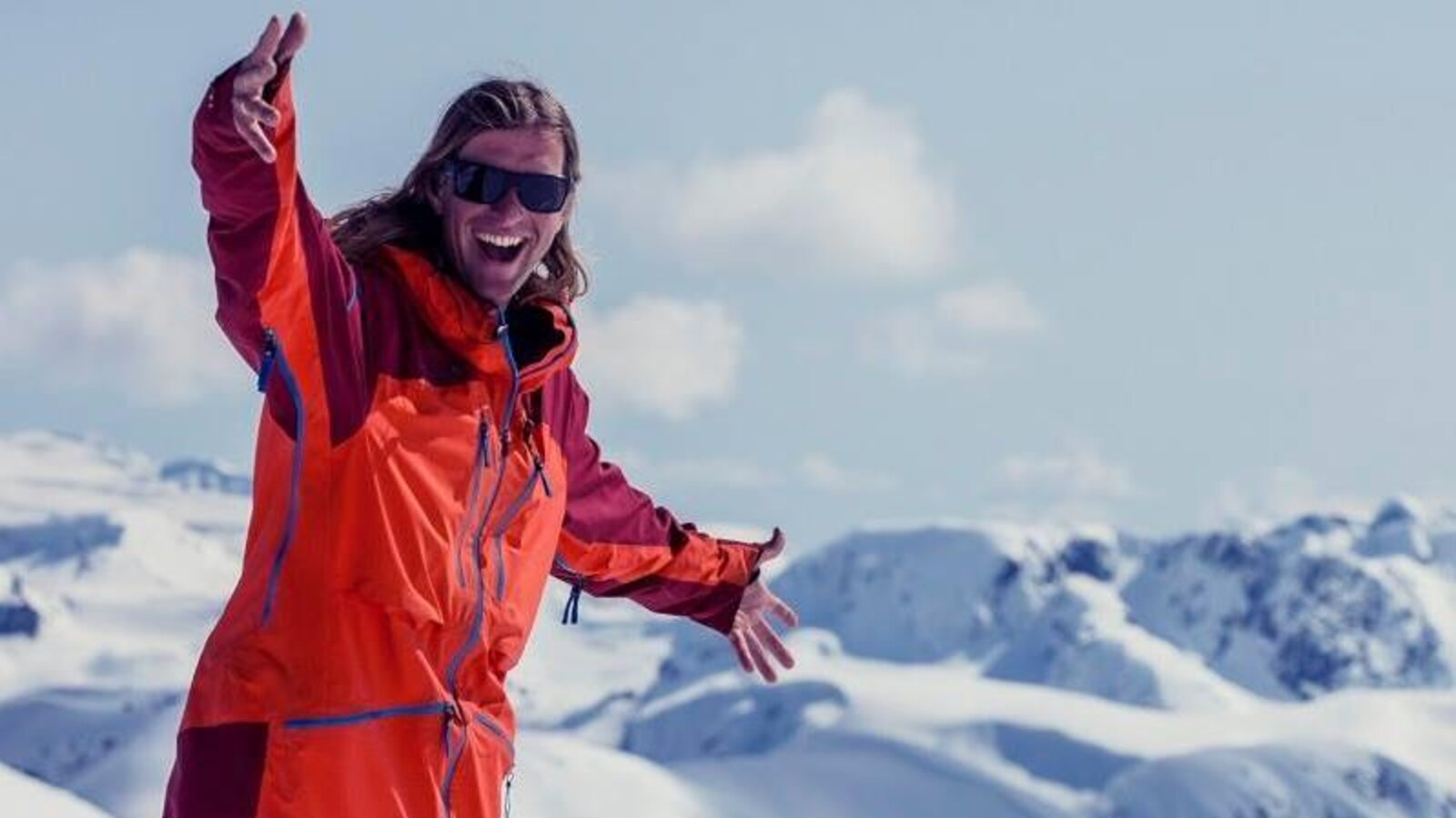 Un homme regarde vers la caméra en souriant, il a un manteau d'hiver, il se trouve sur un sommet de montagne enneigé.