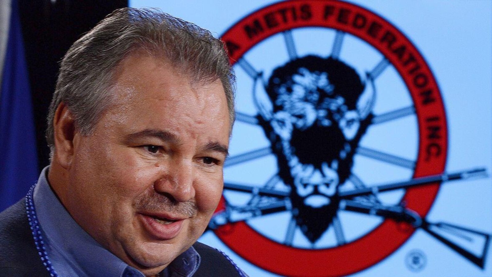 Un homme pose devant le logo de la FMM, sur lequel figurent une tête de buffle et deux carabines.
