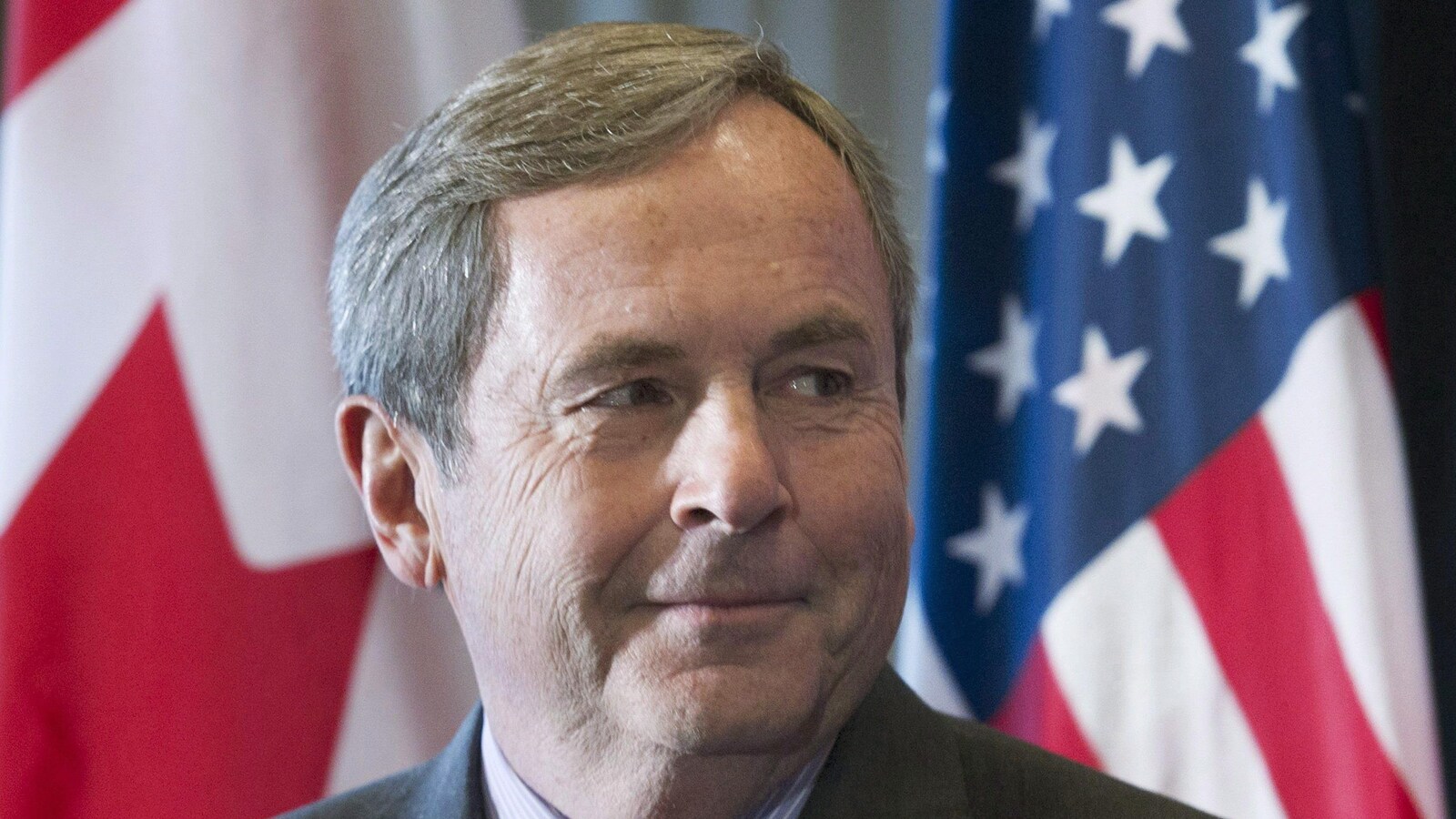 L'ambassadeur se tient devant des drapeaux des États-Unis et du Canada.
