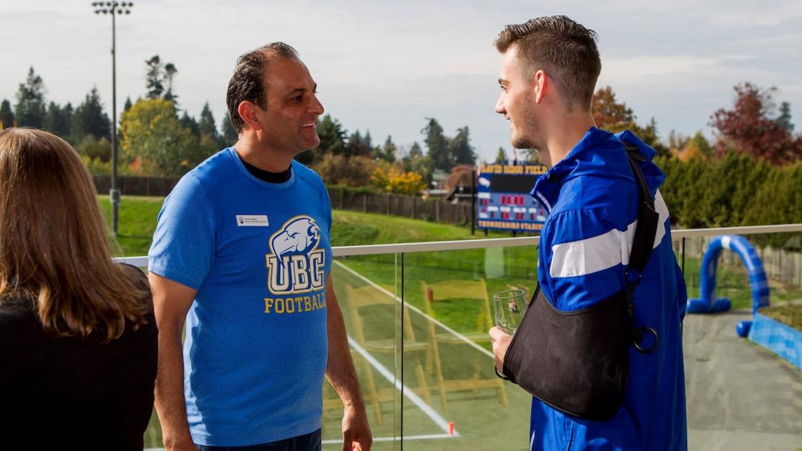 Une homme avec un chandail UBC Football parle avec un joueur de football sur le terrain.