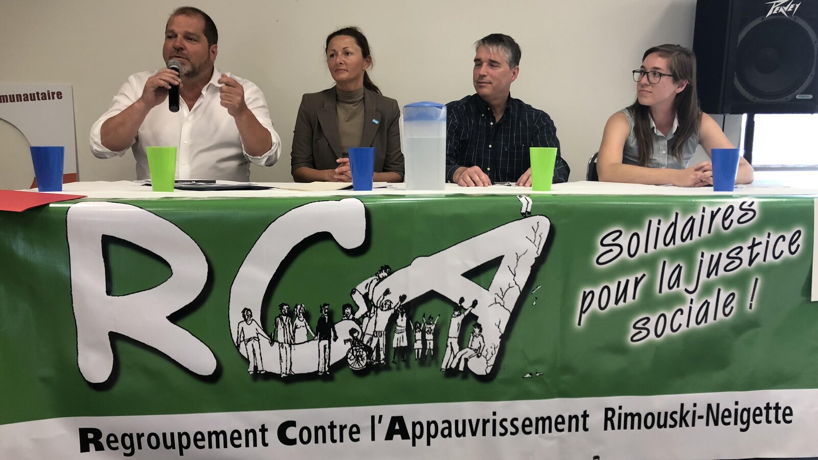 Les quatre candidats sont assis à une table recouverte d'une bannière. Harold Lebel parle au micro.