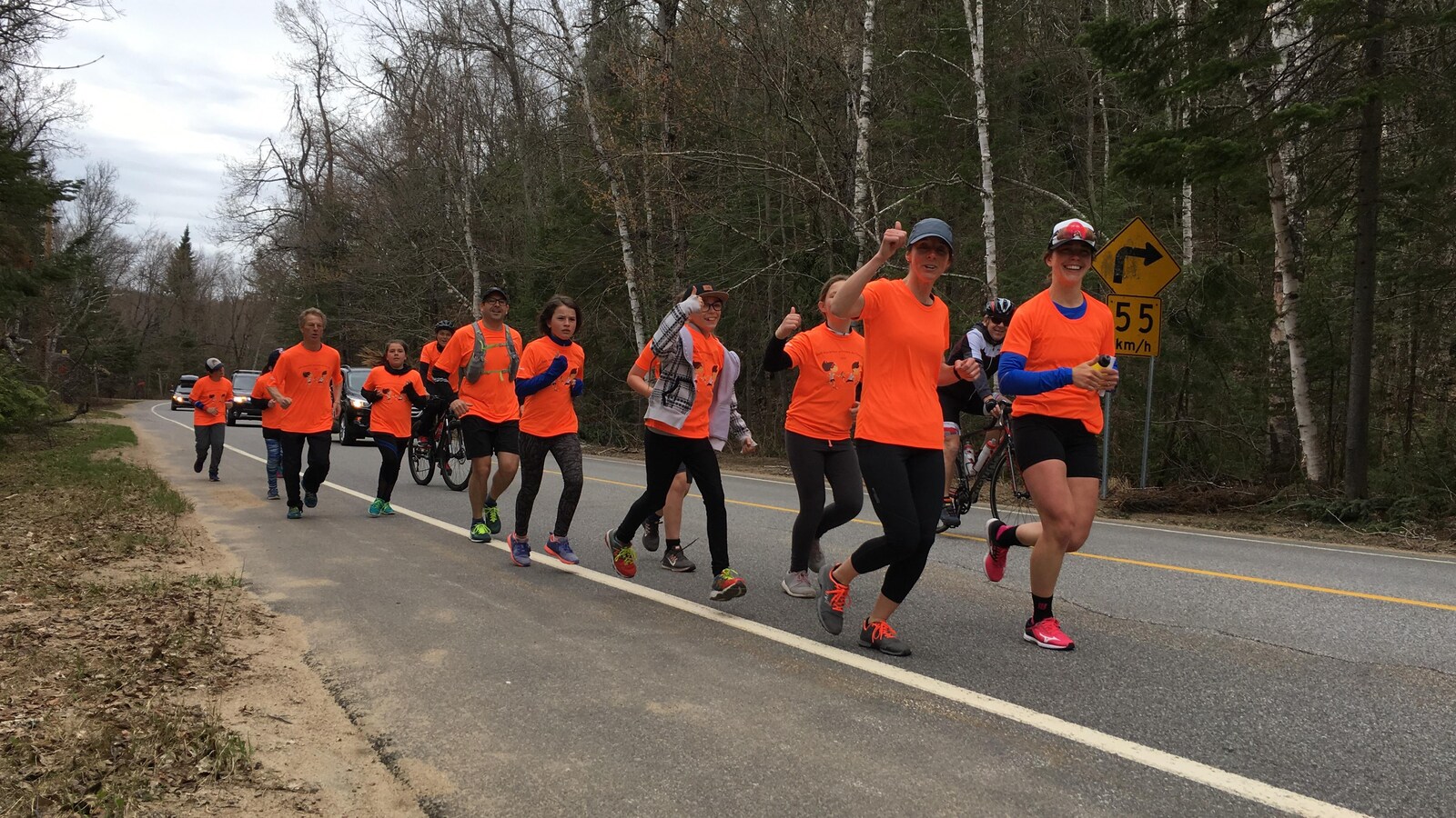 Sur une route de campagne, des adultes et des enfants portant des chandails orange courent en souriant à la caméra.