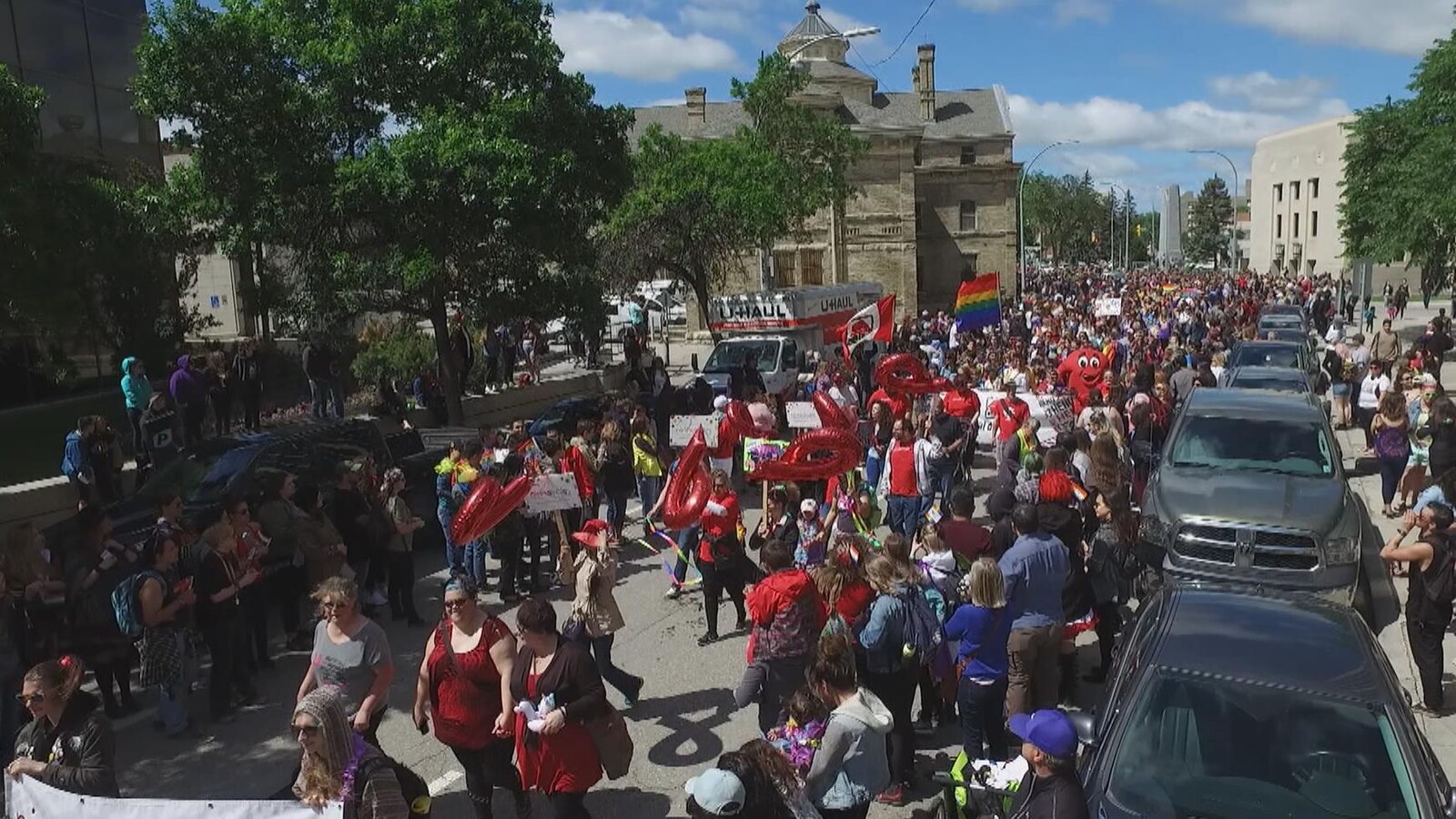 Vue sur le cortège qui défile dans la rue. Les gens portent des vêtements colorés et des pancartes.