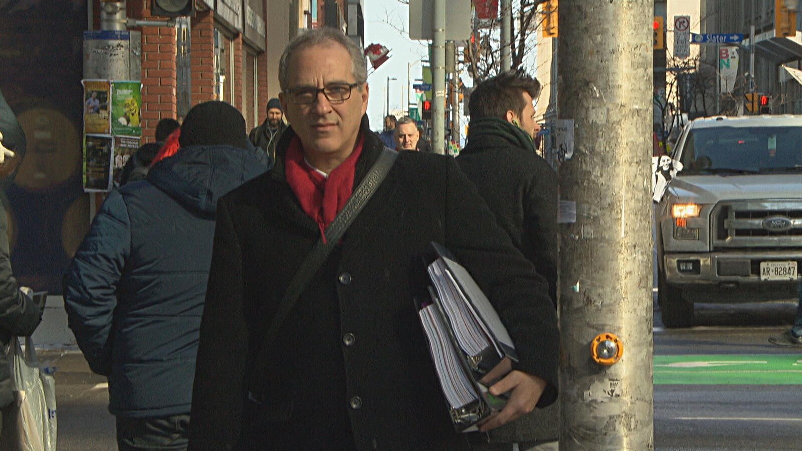 Un homme au coin d'une rue à Ottawa avec deux cartables remplis de feuilles sous les bras.