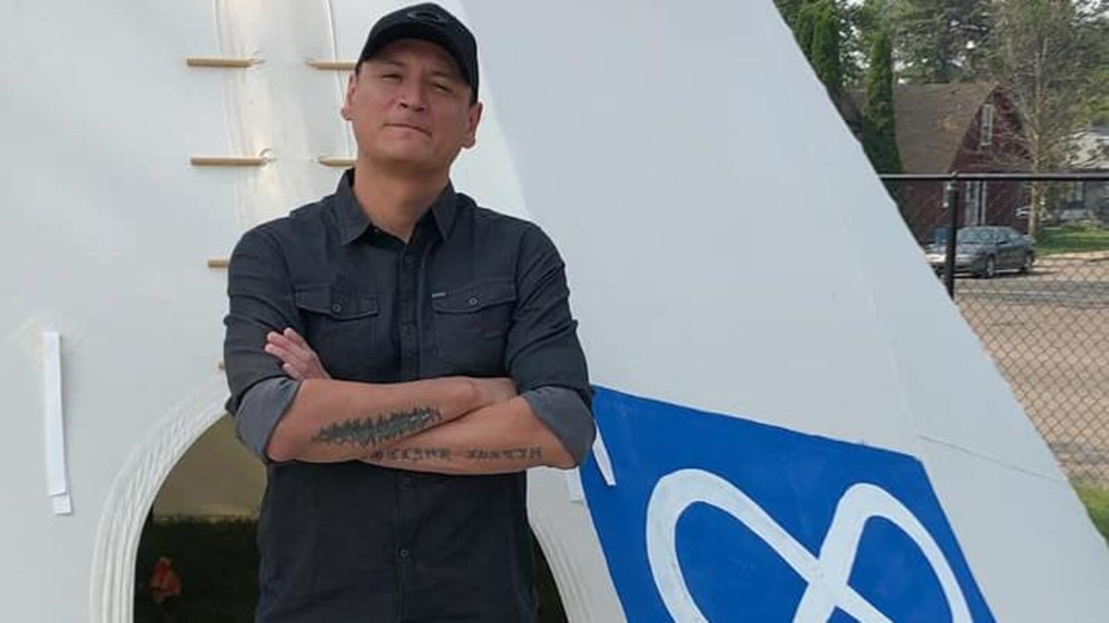 Un homme se tient les bras croisés à l'entrée d'un tipi sur lequel est dessiné le signe métis.