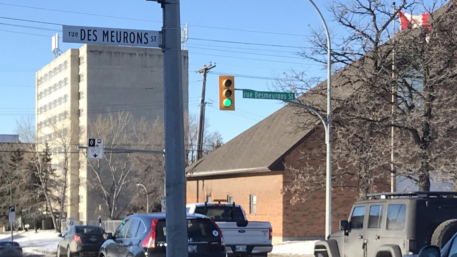 Au bord d'une route, à gauche, un panneau blanc orthographie Des Meurons en deux mots avec une majuscule à Meurons, et à droite un autre panneau vert l'orthographie en un mot.