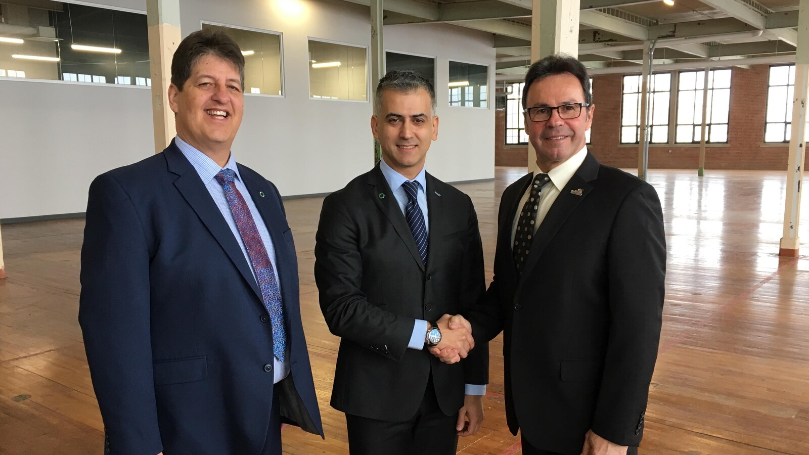 Trois hommes en complet sourient pour la caméra, alors que les deux à droite se serrent la main.