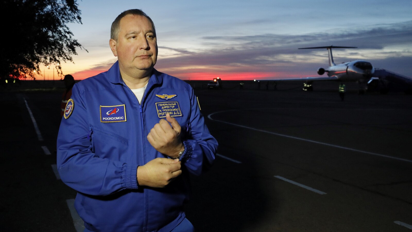 L'homme est sur la piste d'atterrissage. On voit un avion derrière lui et un ciel rosé.