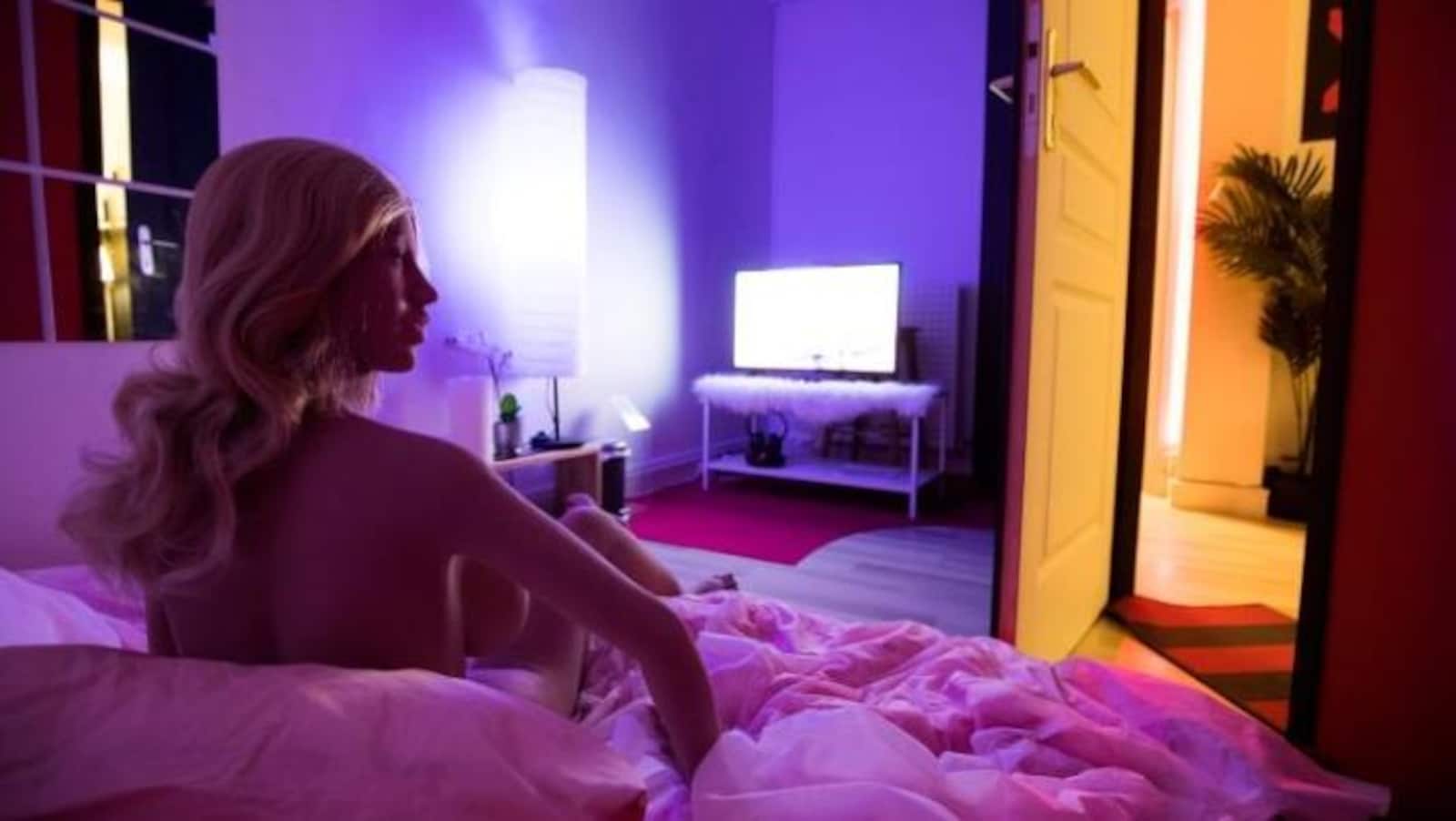 une poupée en silicone nue sur un lit, blonde, regarde la télé