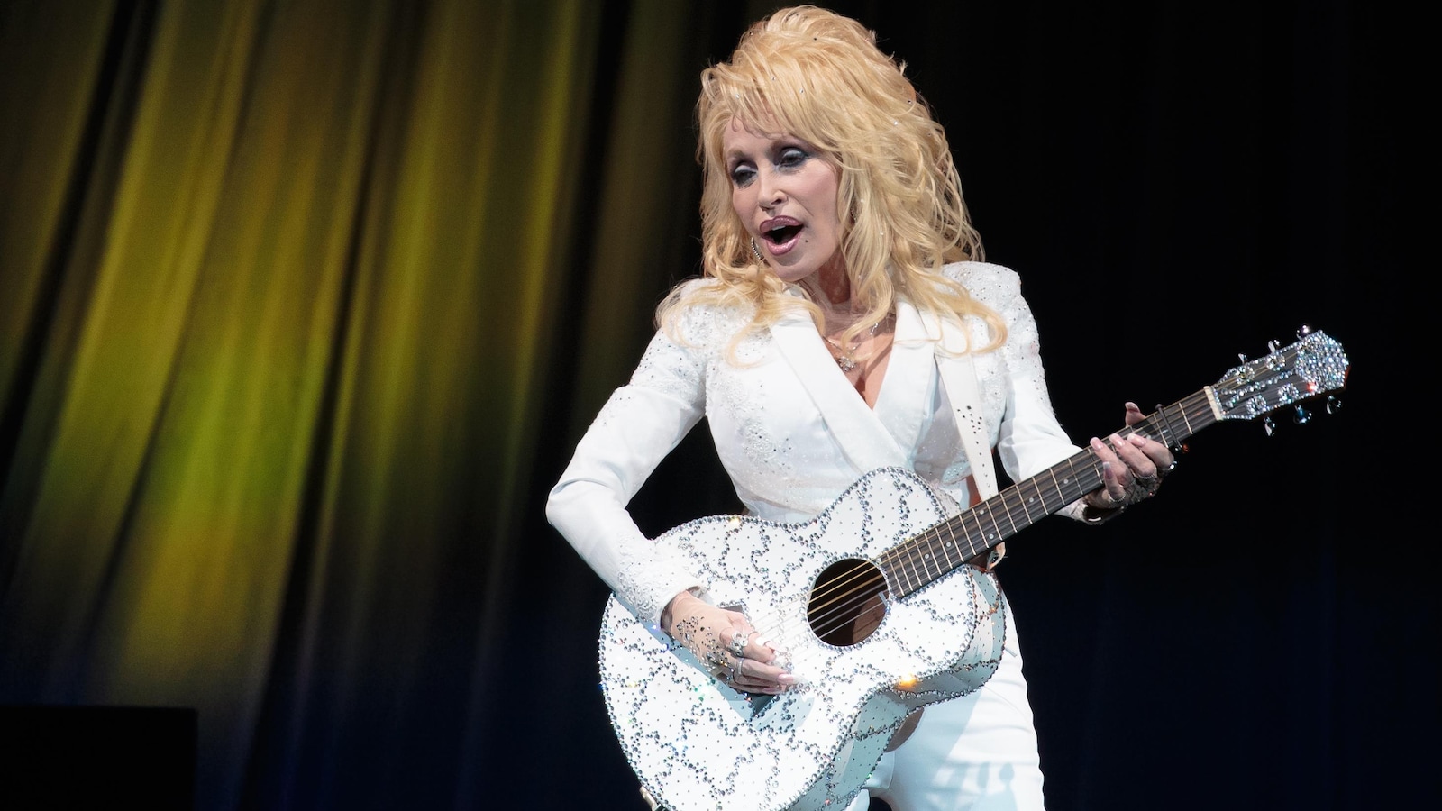 La femme chante sur scène toute habillée de blanc et joue de la guitare.