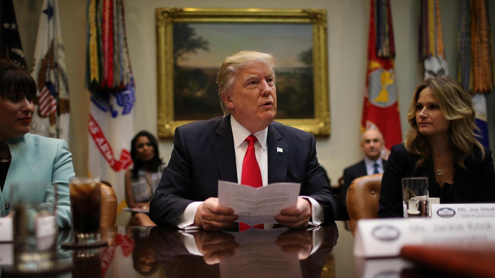 Donald Trump à la Maison-Blanche, assis à une table une feuille à la main et entouré de deux femmes.