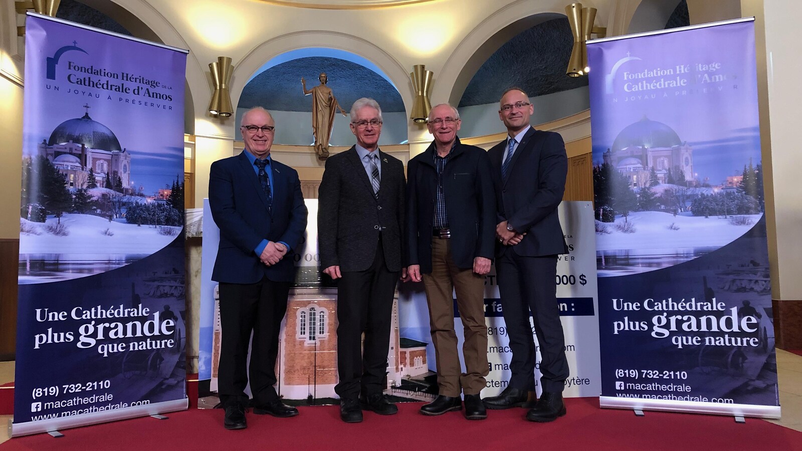 Quatre hommes posent sur l'hôtel d'une cathédrale entre deux affiches de la Fondation Héritage Cathédrale d'Amos.