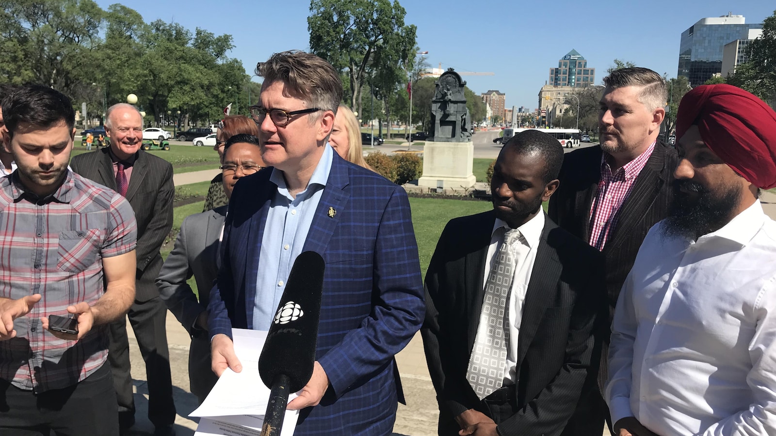 Un homme en veston tenant des documents, entouré de plusieurs personnes diverses, devant un micro tendu par Radio-Canada, à l'extérieur du palais législatif.