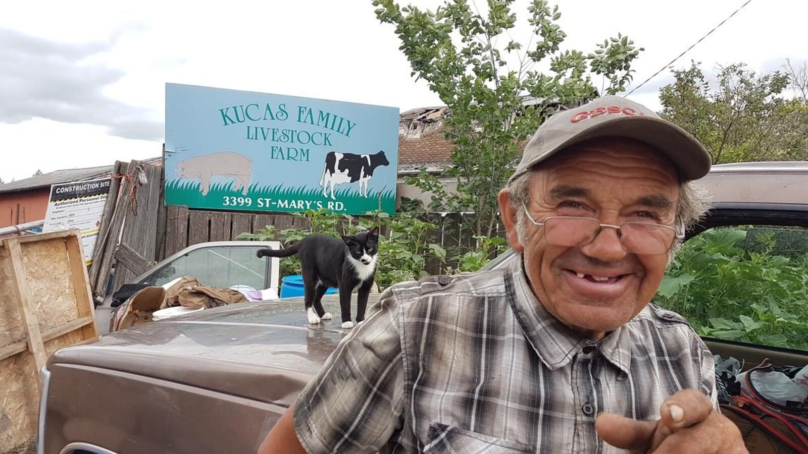 Drago Kucas en gros plan. Derrière lui, un chat sur une vieille voiture et derrière la voiture de multiples objets empilés.