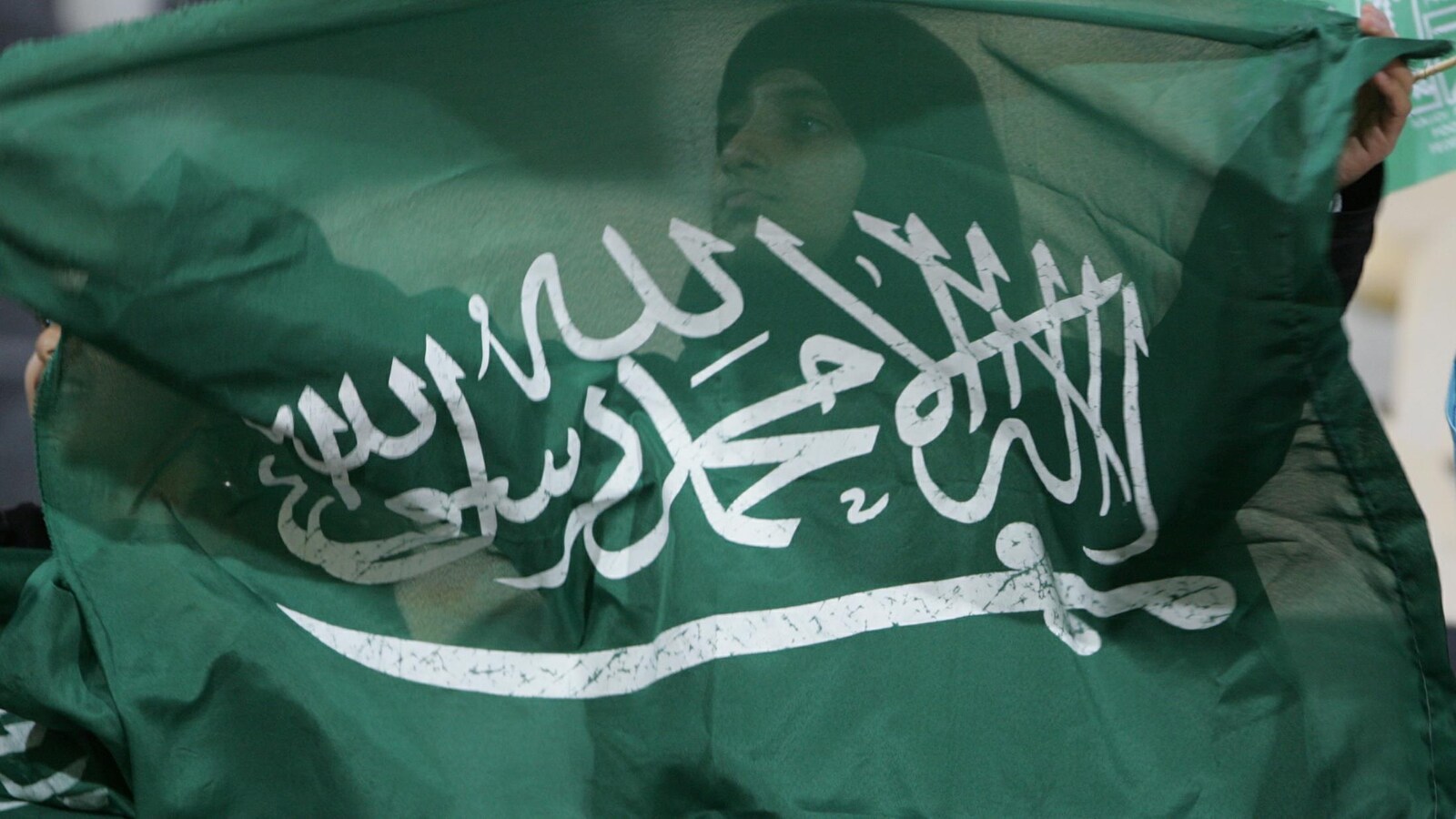 Une femme voilée tient un drapeau de l'Arabie saoudite dans ses mains devant elle. On la voit en transparence à travers le tissus du drapeau.