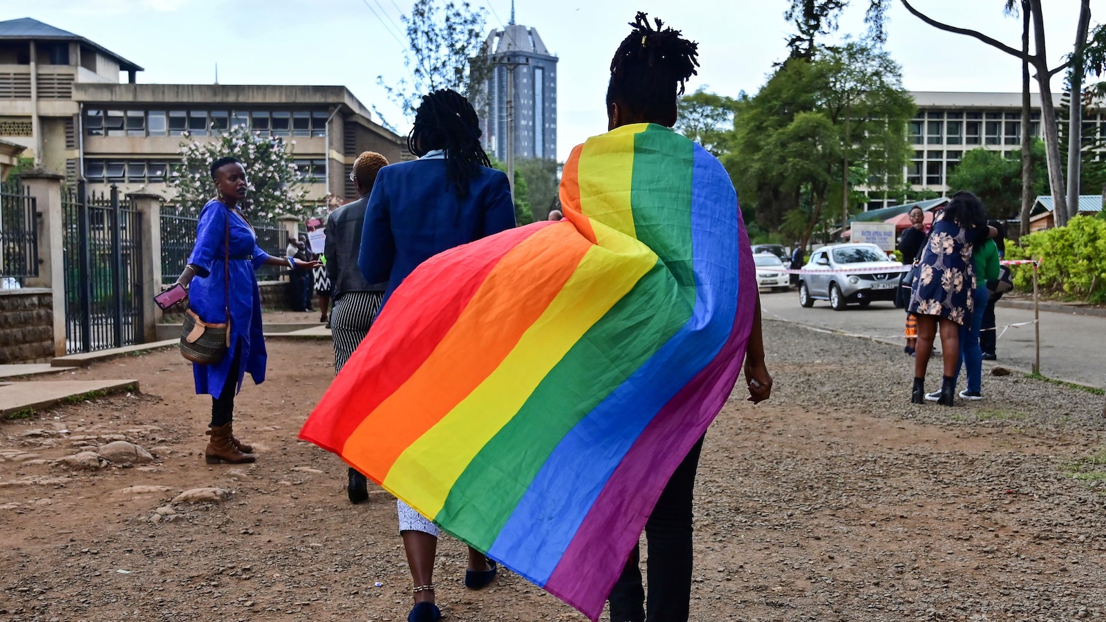 Une personne kenyane, que l'on voit de dos, porte un drapeau arc-en-ciel en guise de cap, et marche sur la voie publique, où se trouvent d'autres personnes.