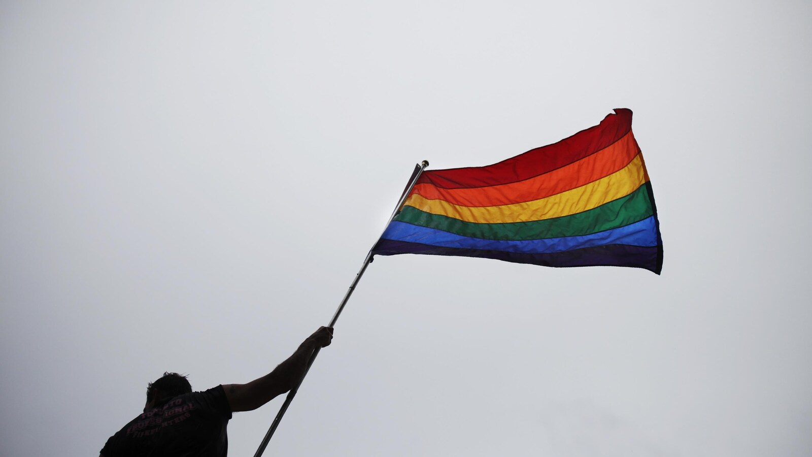 Un homme tient dans sa main droite un drapeau ayant aux couleurs de l'arc-en-ciel. La photo a été prise en contre-plongée, c'est-à-dire que l'on voit de plus bas l'homme et le drapeau, comme si on levait la tête pour les regarder.