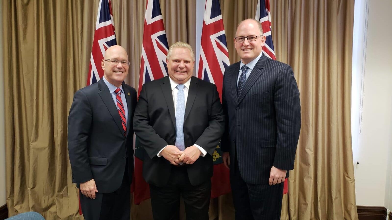 Trois hommes regardent la caméra devant quatre drapeaux de l'Ontario.