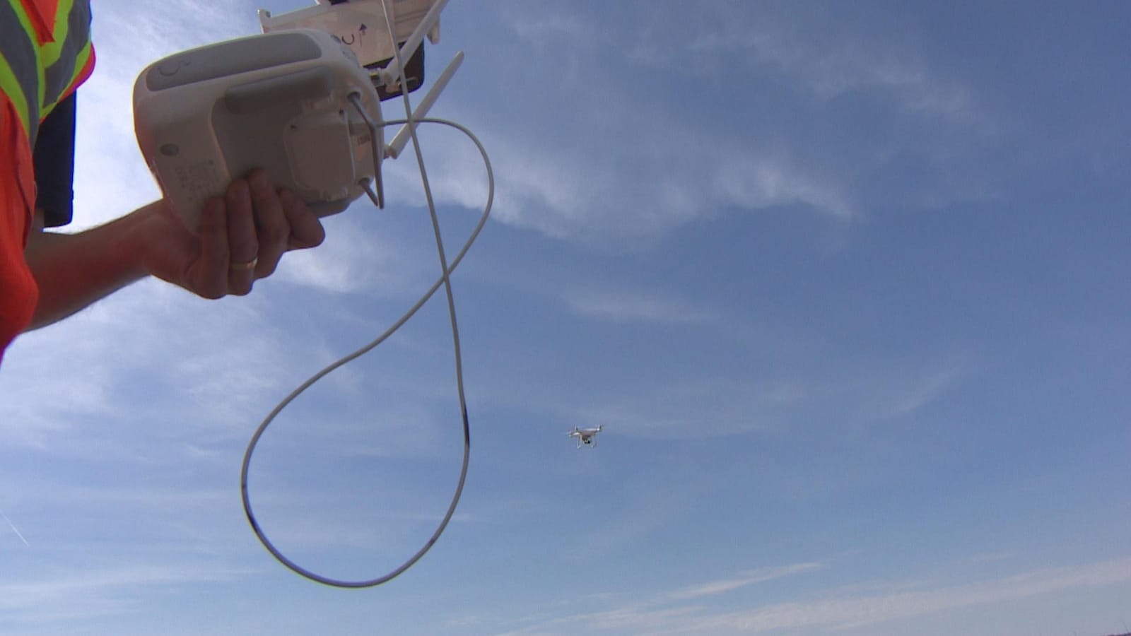 Au premier plan à gauche : les mains d'un pilote de drone manipulent une télécommande. Au fond, en arrière-plan : un drone blanc vole dans le ciel.