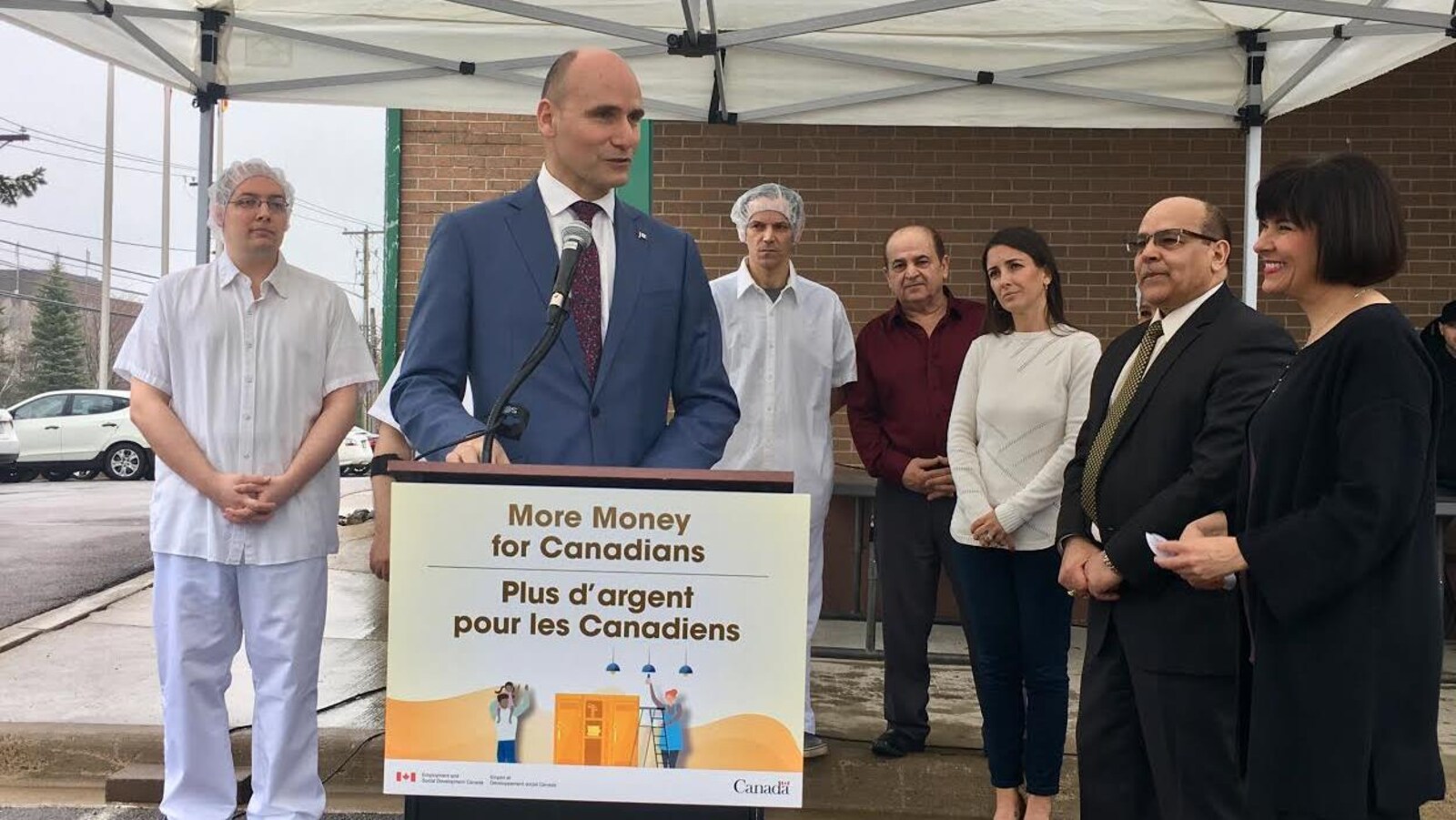 Le ministre Duclos donne une conférence de presse devant la boulangerie, il est accompagné de travailleurs et d'autres personnalités.