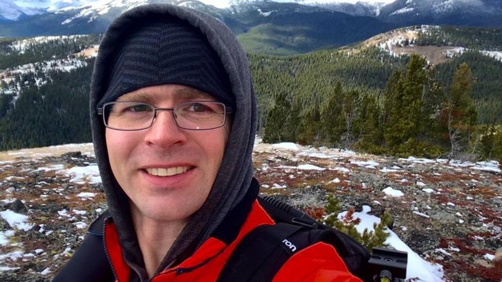 Plan serré du visage de Dwayne Schnell habillé pour la randonnée avec en arrière-plan des montagnes et une forêt en hiver.