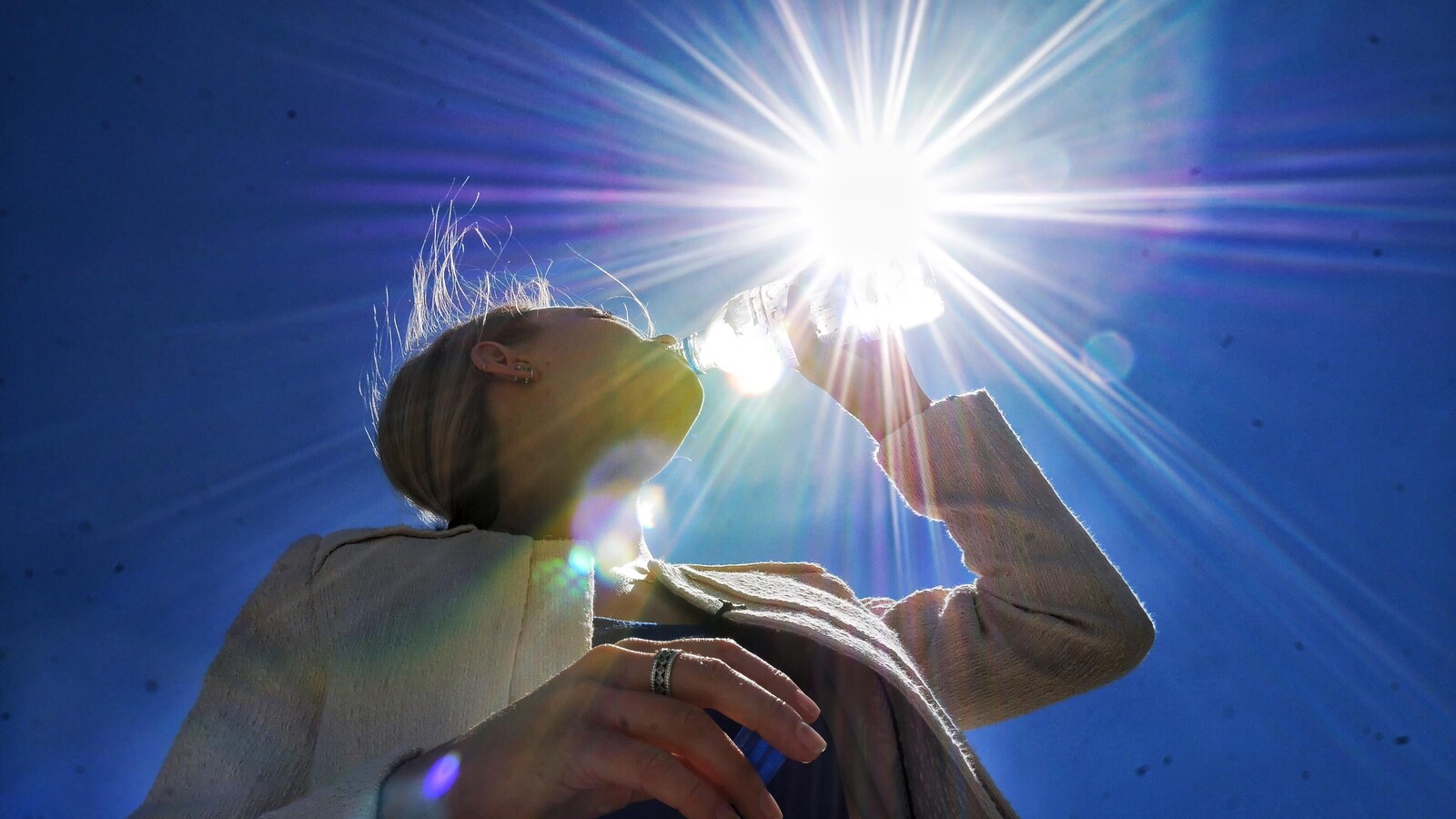 Contre-plongée d'une femme qui porte une bouteille d'eau à ses lèvres, avec le soleil qui plombe en arrière-plan.