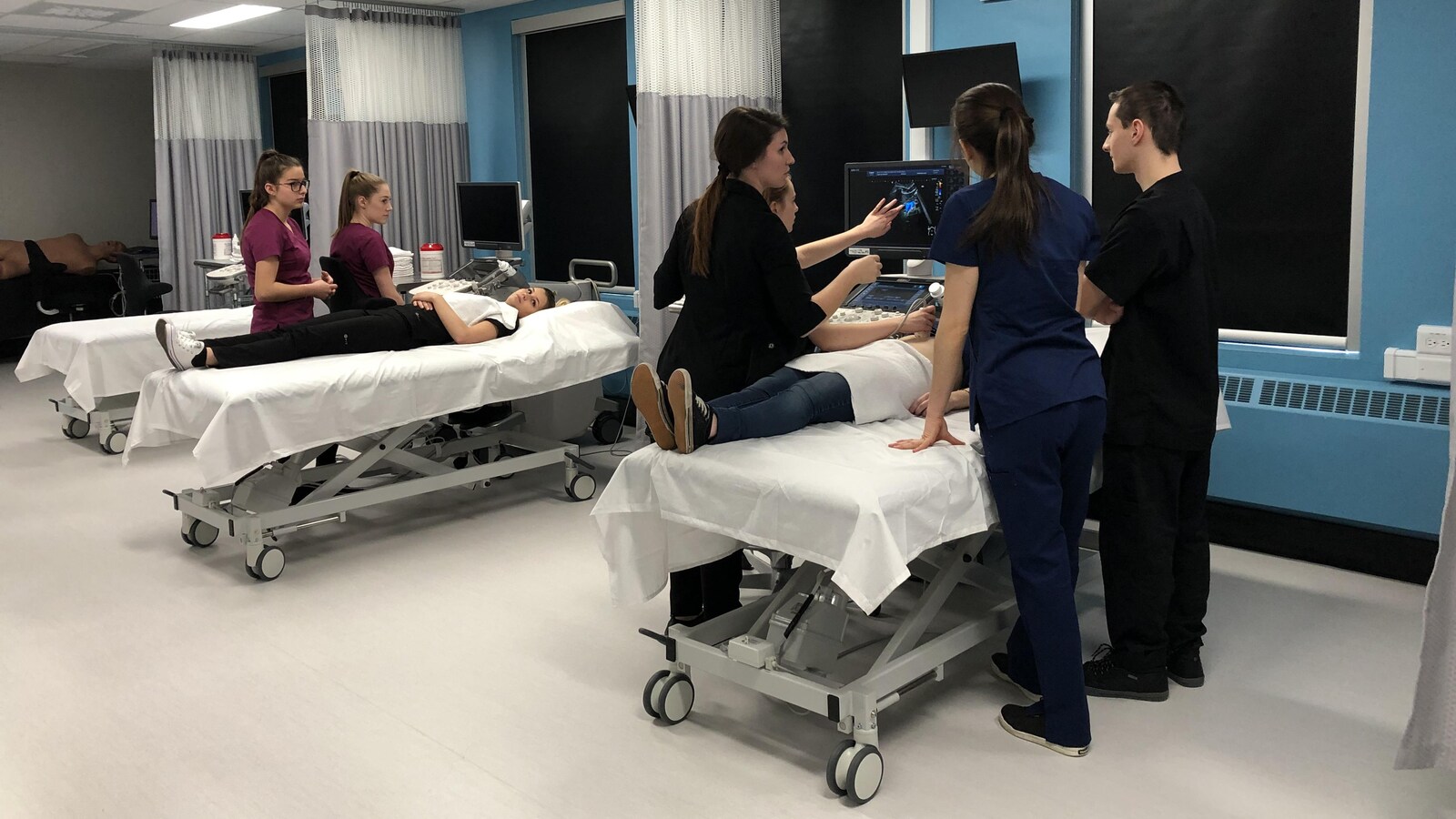 5 étudiants, accompagnés de leurs enseignants, font un test d'échographie sur deux de leurs collègues, étendus sur un lit d'hôpital.