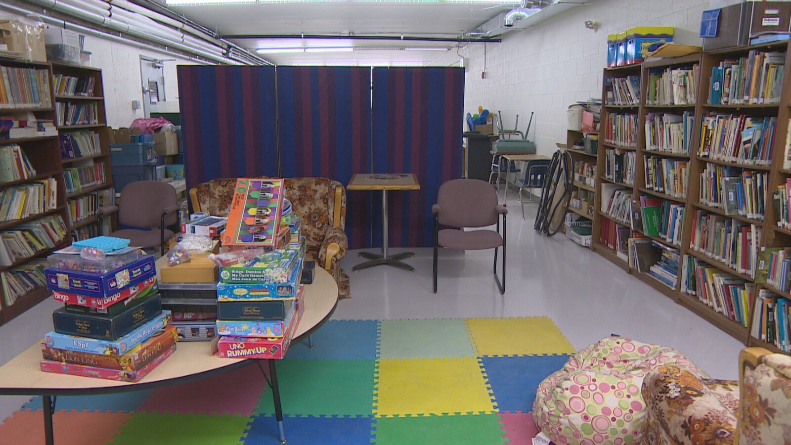 Une salle sans fenêtre autour de laquelle se trouvent plusieurs étagères remplies de livres. Des jeux de société sont empilés sur une table basse.