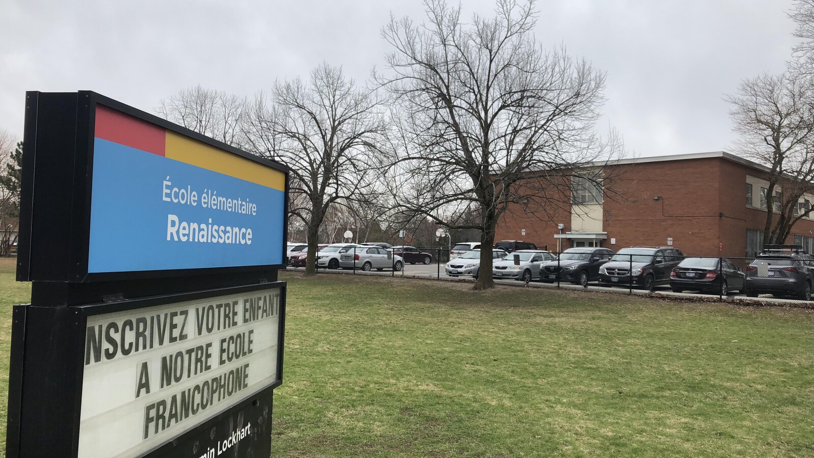 Une pancarte sur laquelle est écrit "inscrivez votre enfant à notre école francophone", avec un bâtiment derrière.