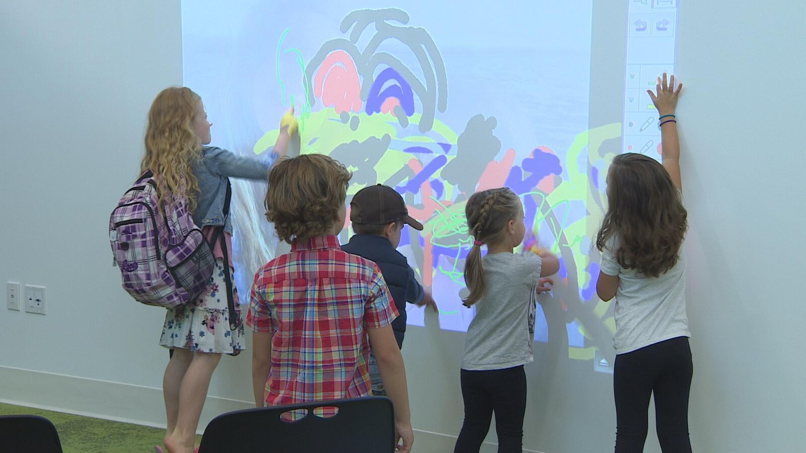 Des enfants debout devant un mur blanc sur lequel est projeté un carré de lumière où ils font des dessins de couleur.