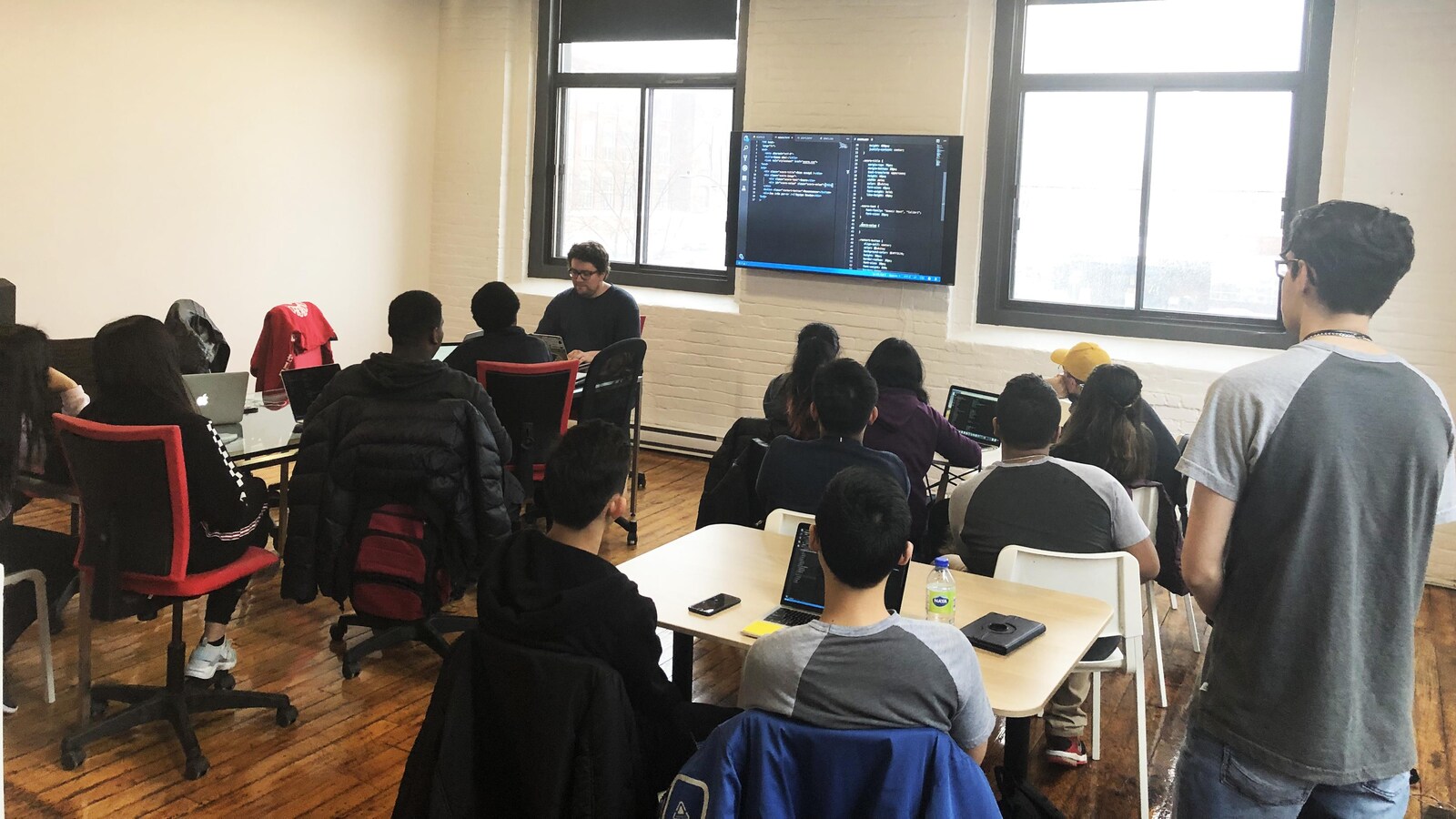 Une photo montrant une salle de classe. Une douzaine d'adolescents sont assis, dos tourné à la caméra, et regardent un téléviseur sur lequel est affiché du code informatique.