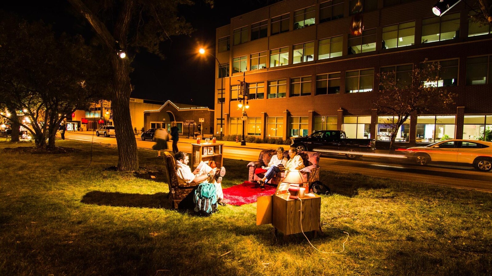 Assis sur un canapé et un fauteuil, des lecteurs lisent dans un parc. C'est la nuit, des lampes éclairent le parc.