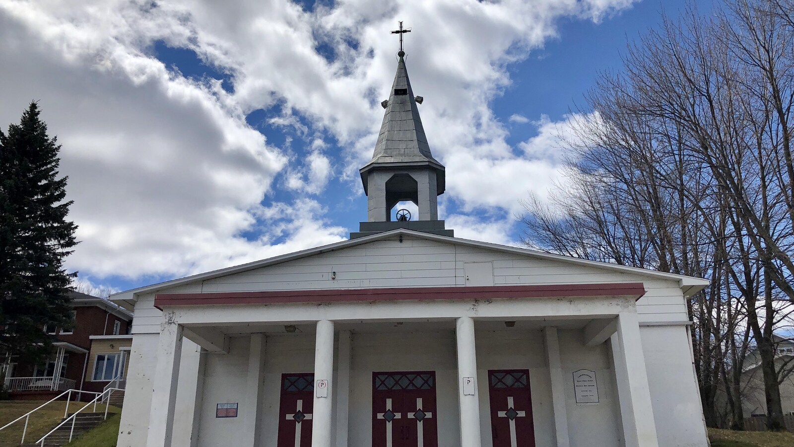 L'église est blanche et il y a des portes rouges. On peut voir au-dessus le clocher gris avec une croix.