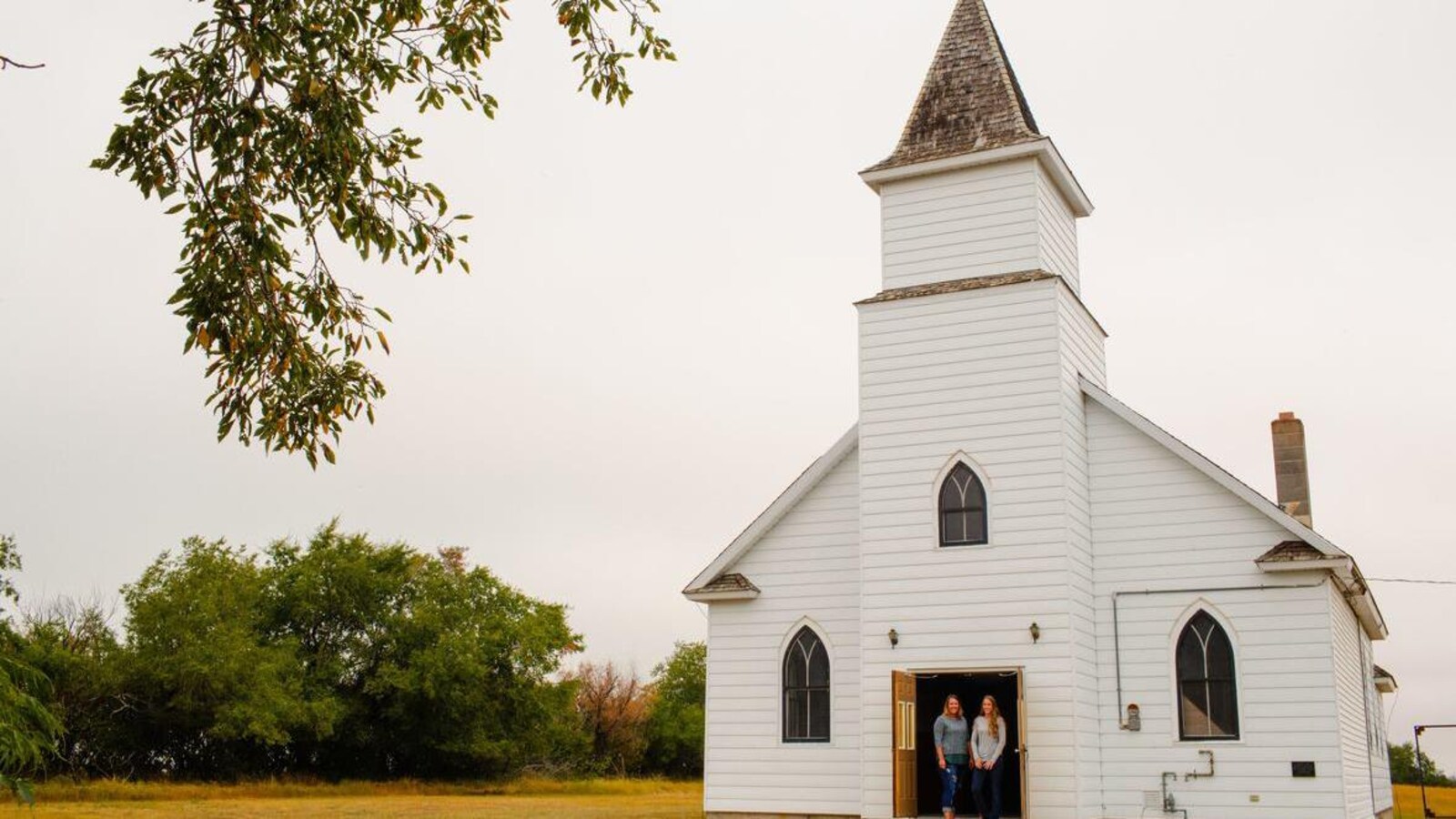 Deux femmes sur le perron d'une petite église blanche, sur un terrain vague.