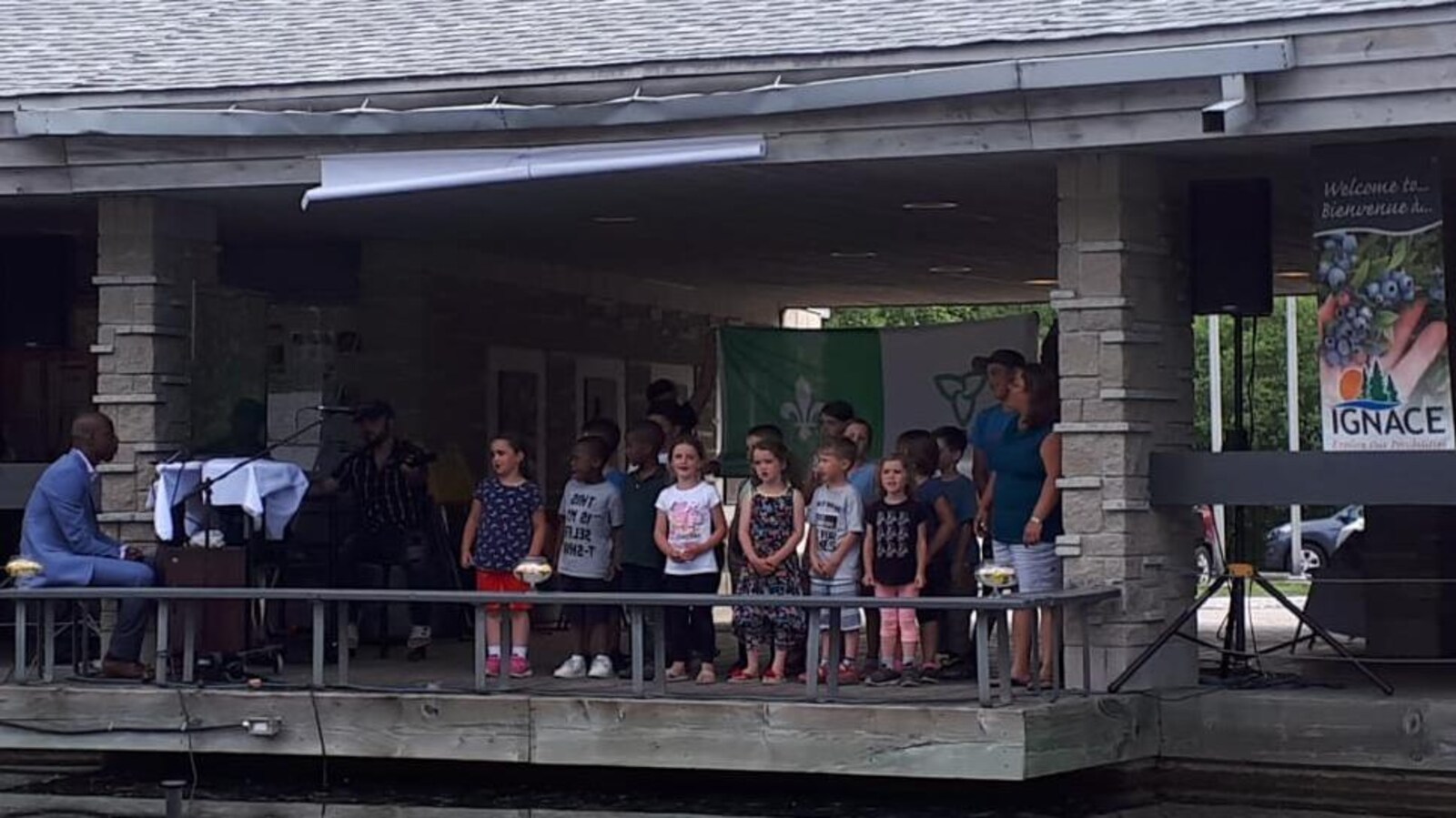 Un groupe d'enfant en train de chanter, accompagné par un pianiste, devant le drapeau franco-ontarien.