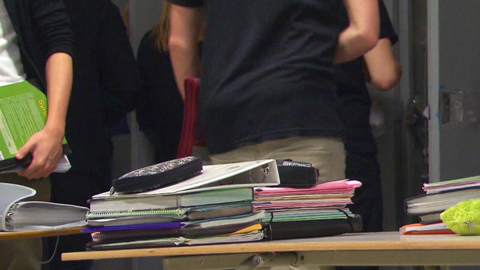 Des cartables et des livres sont posés sur une table dans une classe avec des élèves autour dont on ne voit pas le visage