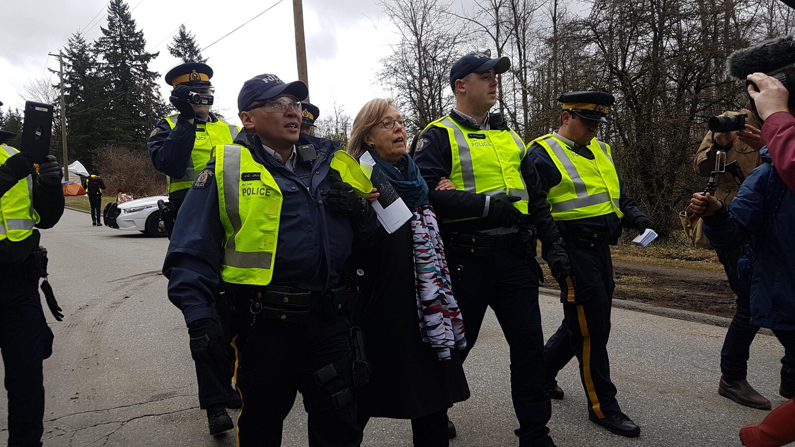 Une dame au bras de deux policiers et entourée de trois autres est escortée dans une rue, près d'une forêt, face aux caméras de télévision.