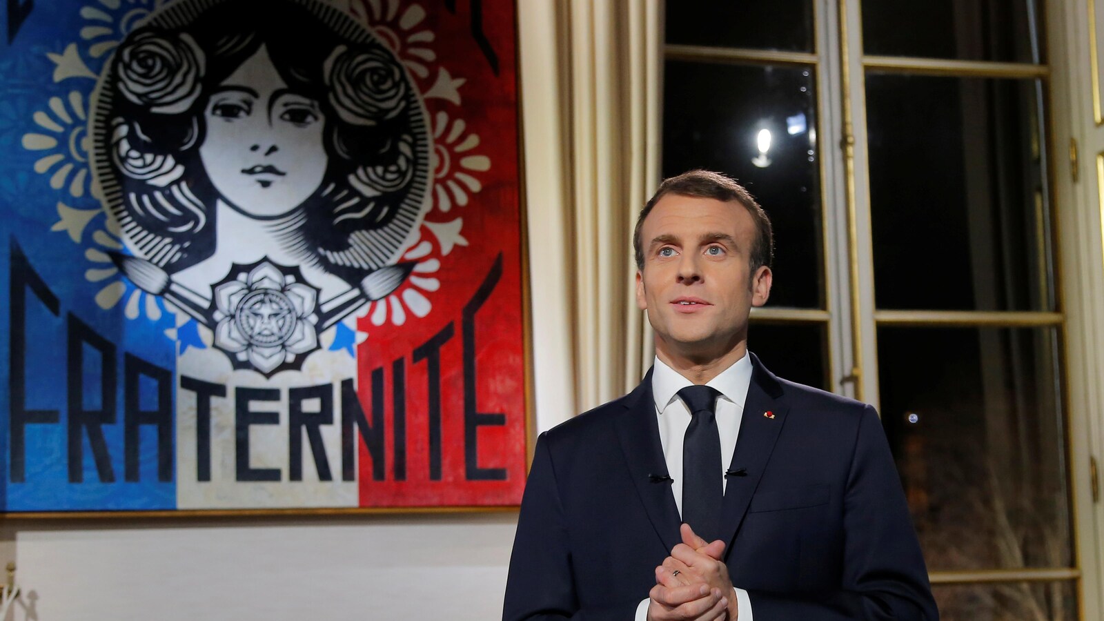 Emmanuel Macron, dans un bureau de l'Élysée, regarde devant lui, les mains jointes, devant une affiche représentant le visage d'une femme au-dessus du mot «fraternité» en lettres majuscules.