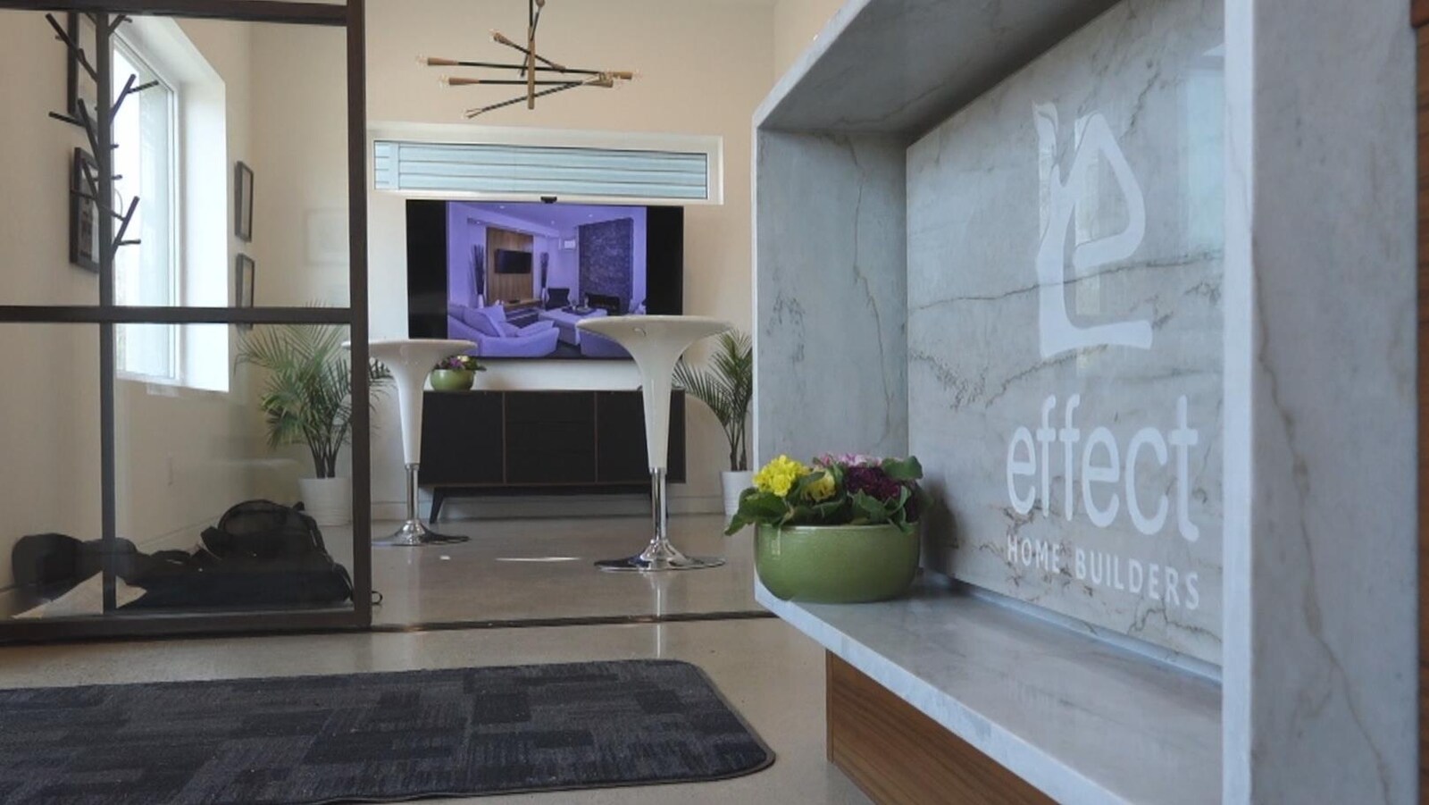 L'intérieur de l'immeuble. Au premier plan, il y a une enseigne en marbre et un pot de fleur. On voit des chaises blanches une télévision et des plantes à l'arrière.